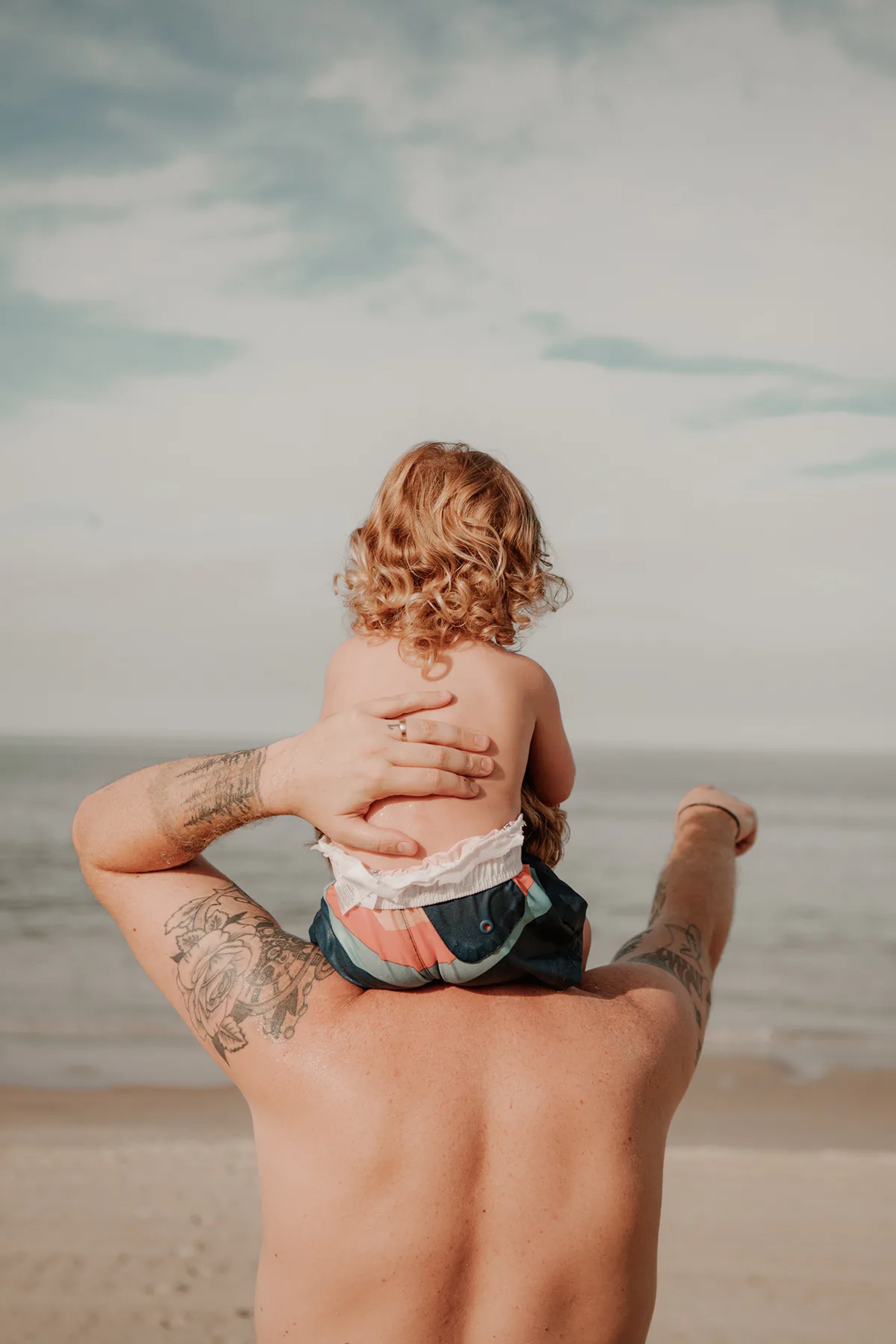 Young child riding on an adult's shoulders while facing the ocean.