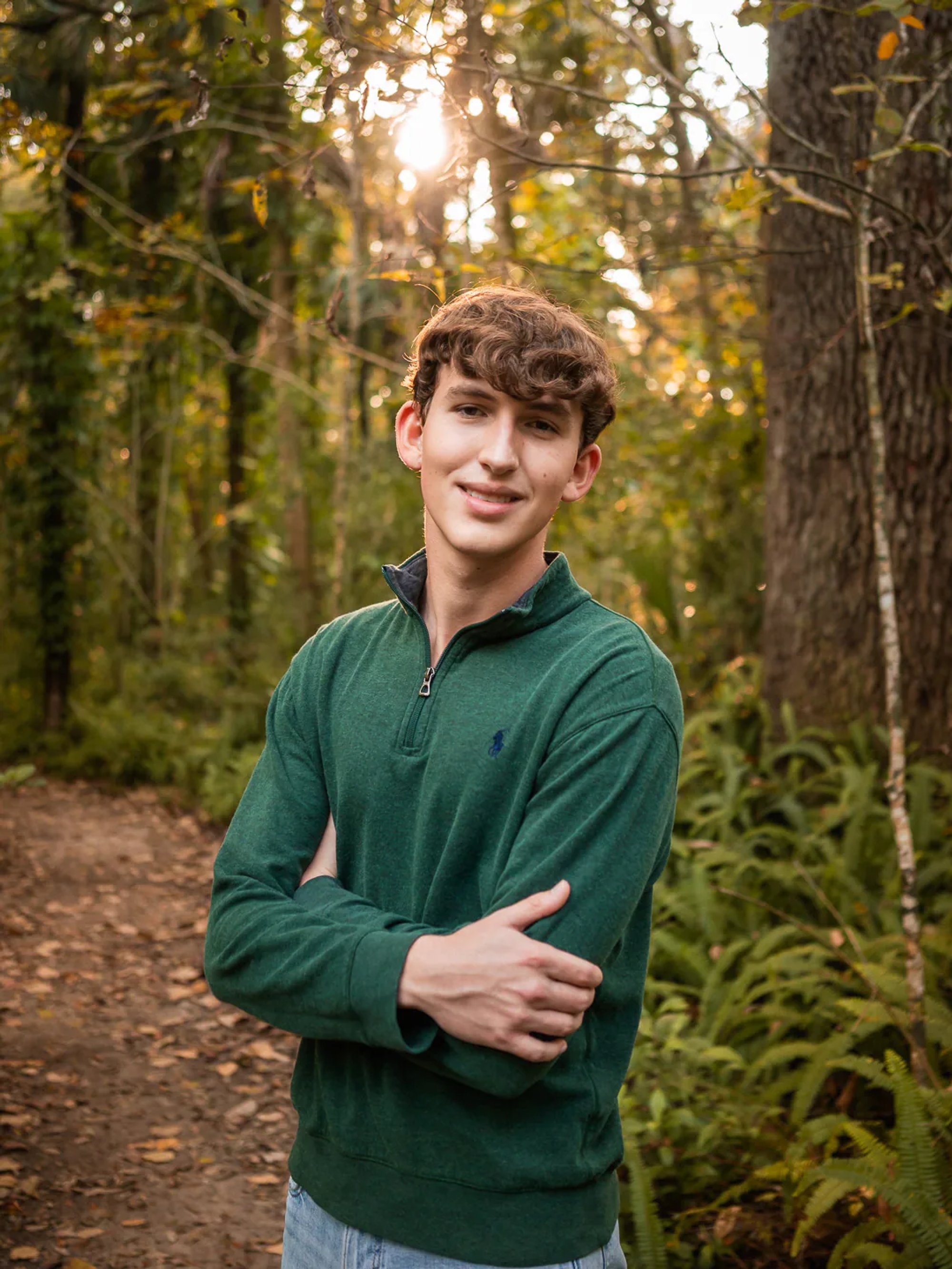 Young man in a green shirt with his arms crossed on a wooded path.
