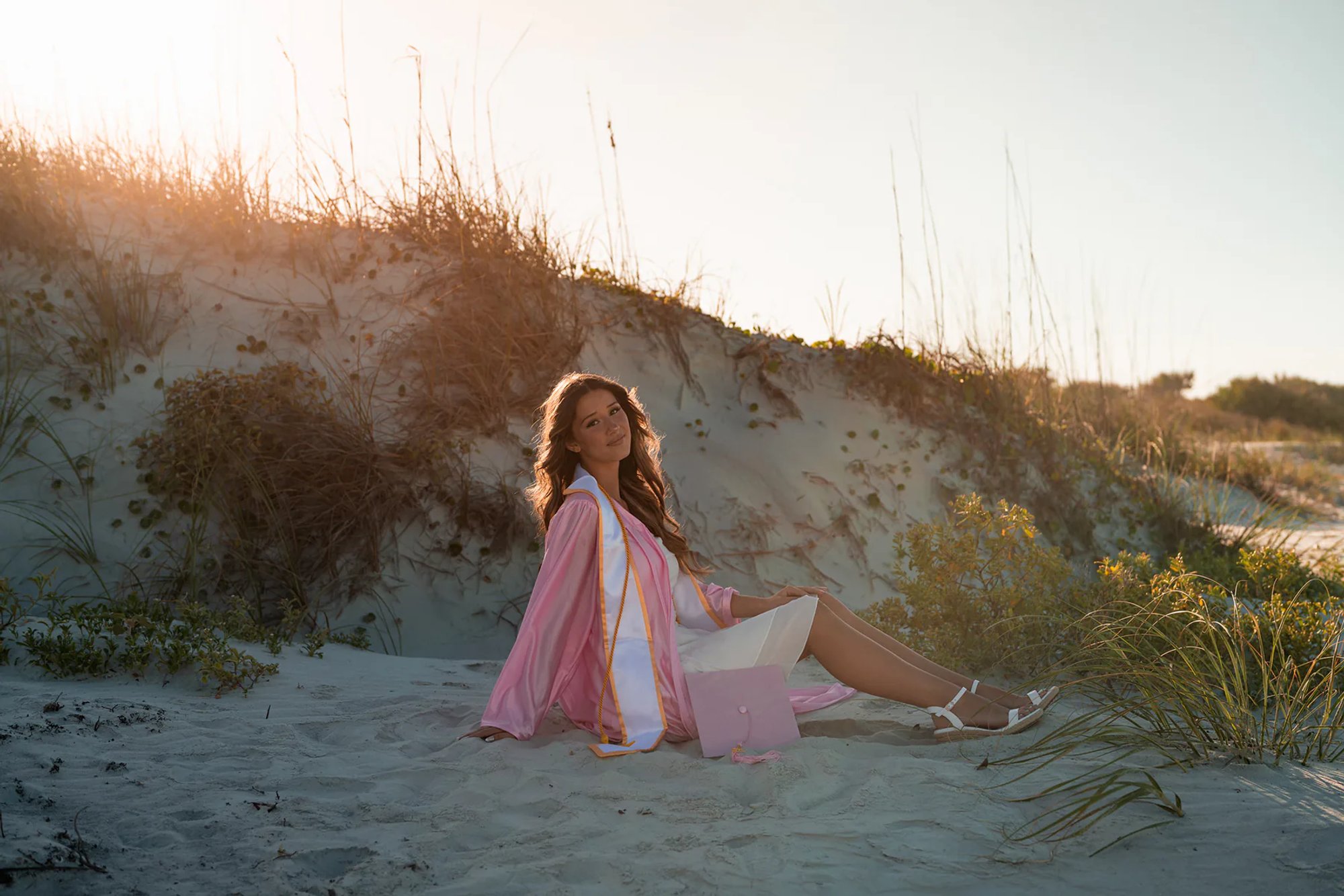 Young woman in a pink wrap and white dress seated beside a sandy dune.