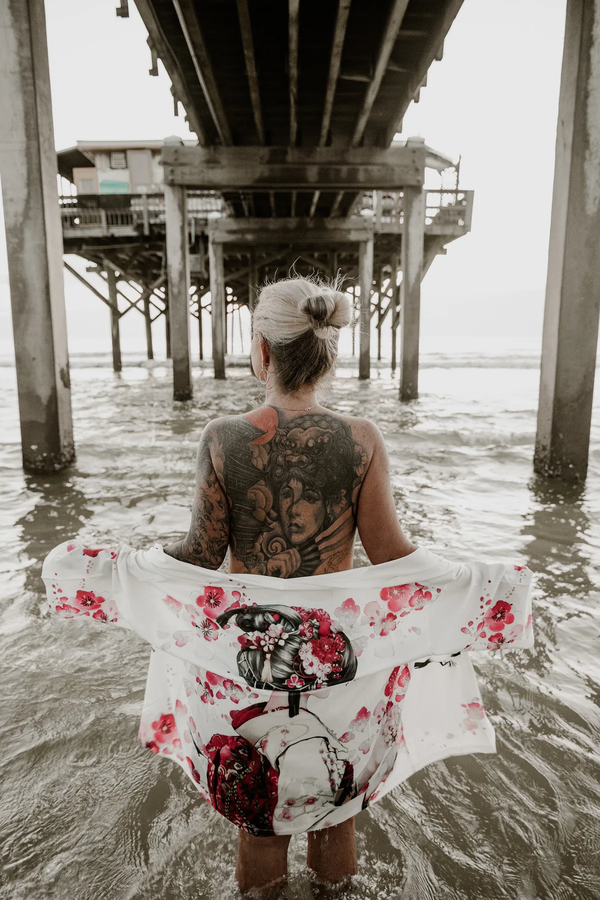 Tattooed woman holding a floral wrap while standing in the water beneath a pier.