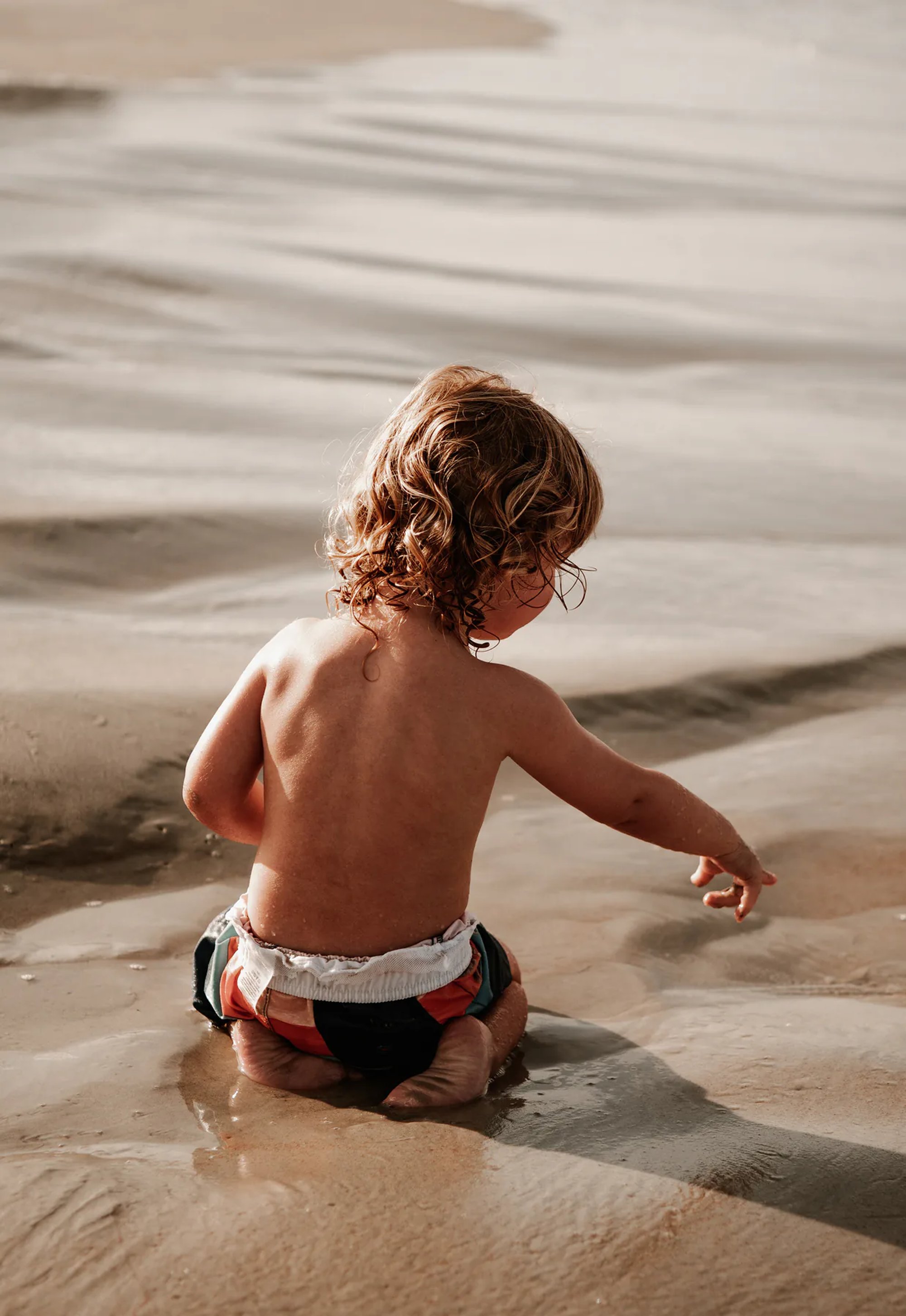 Toddler sitting at the edge of shallow beach water.