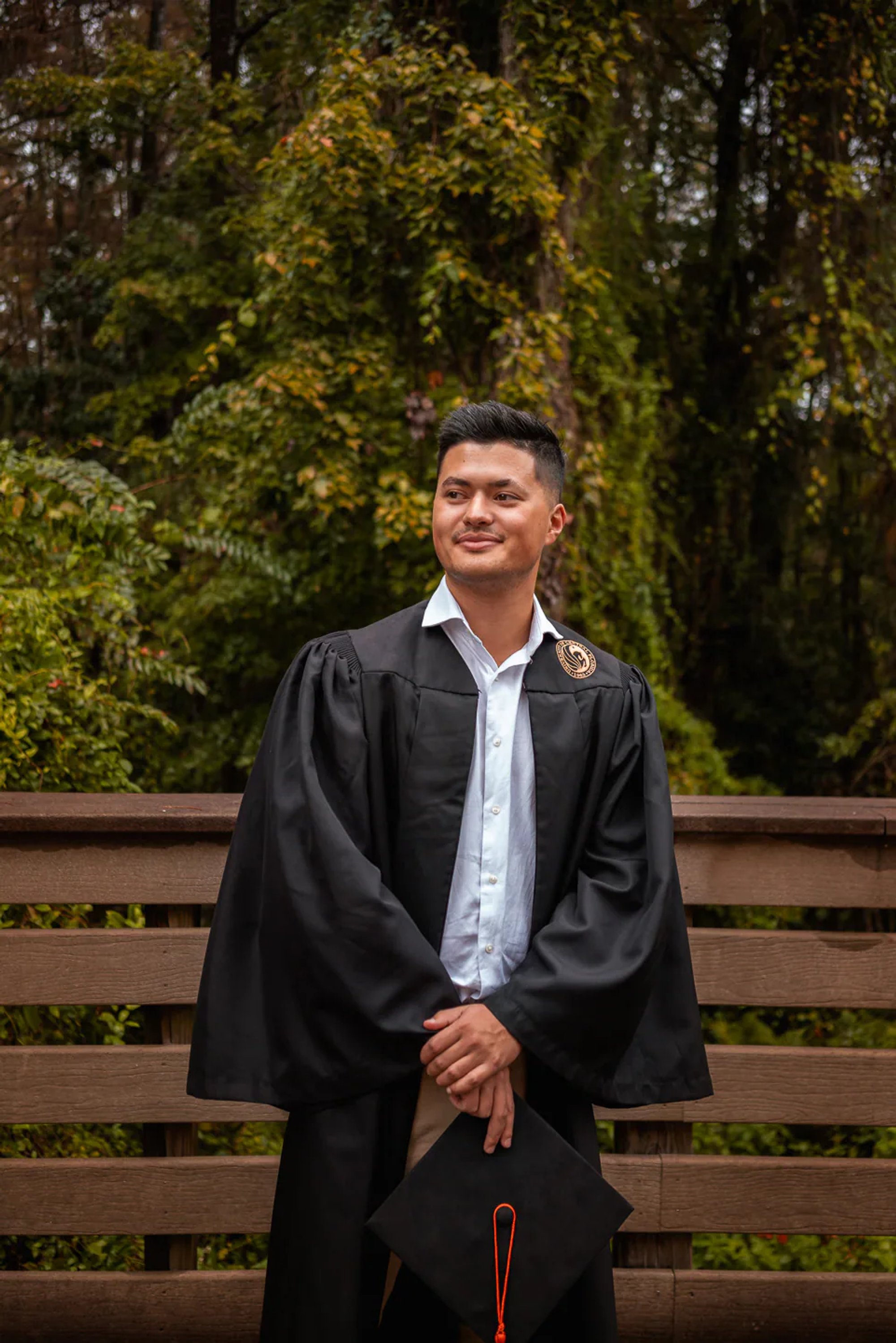 Male graduate in a black gown standing beside a wooden railing.