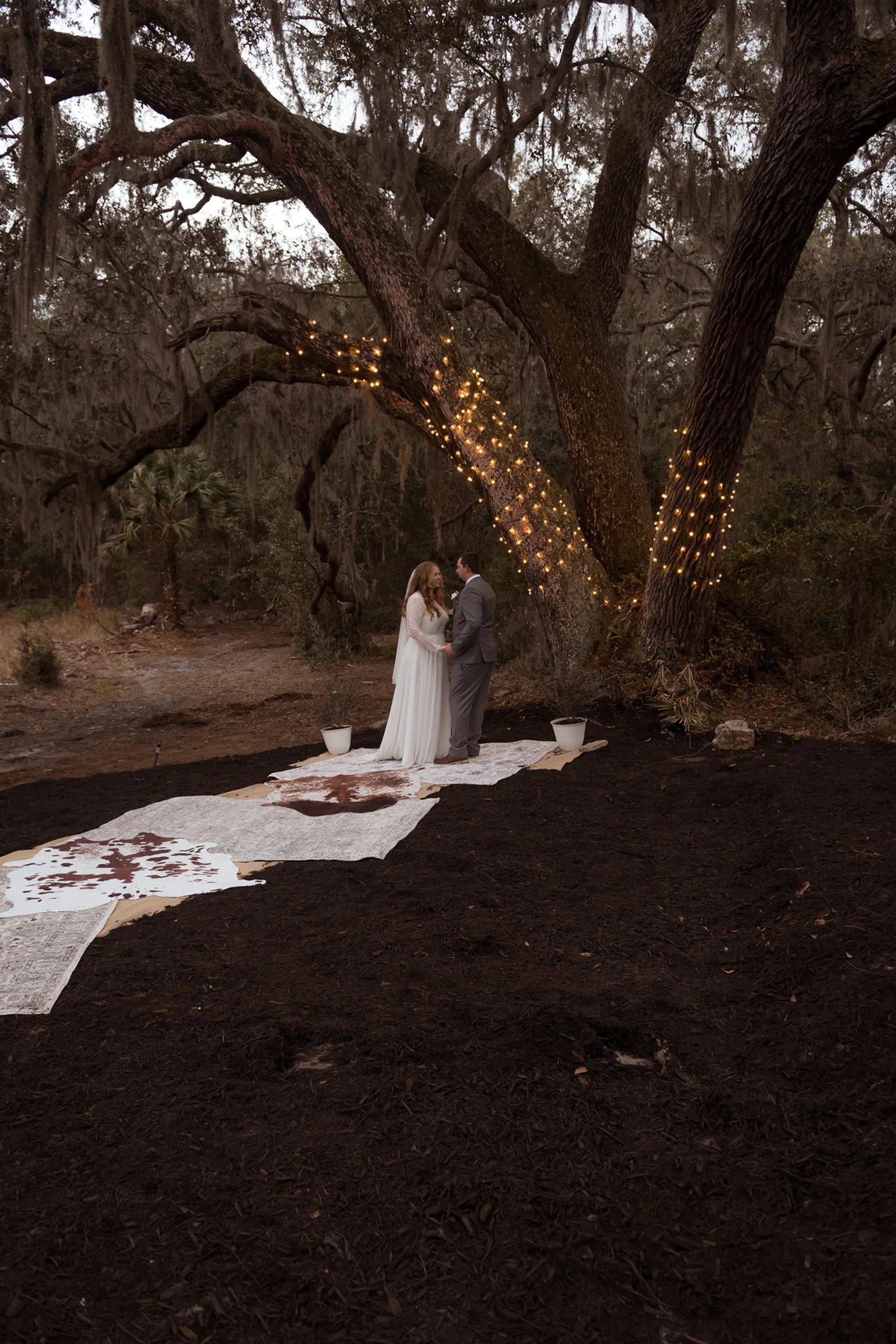 Bride and groom standing at the end of an outdoor aisle beneath sprawling oak trees.