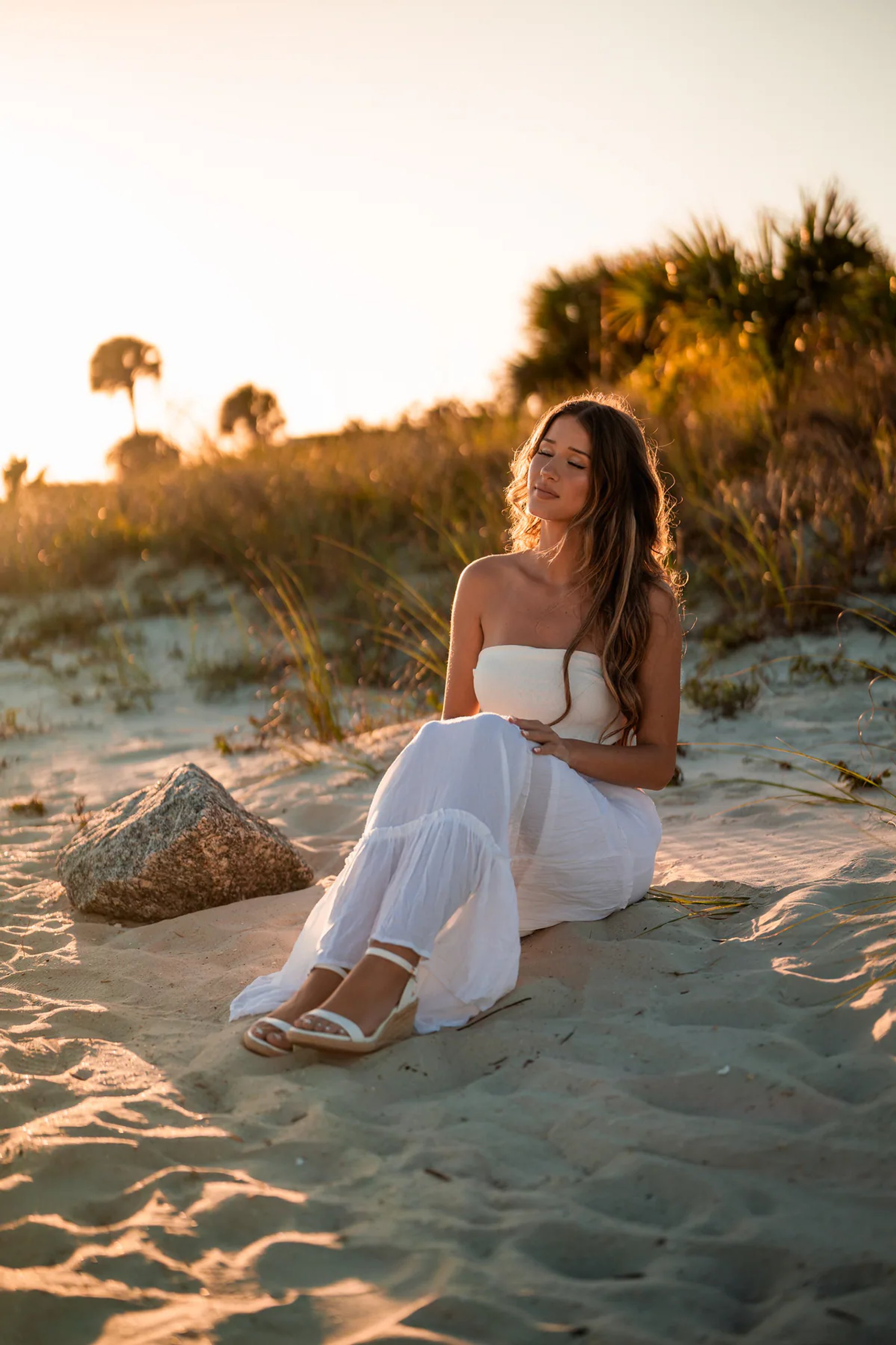 Young woman in a white dress seated on the sand beside a dune.
