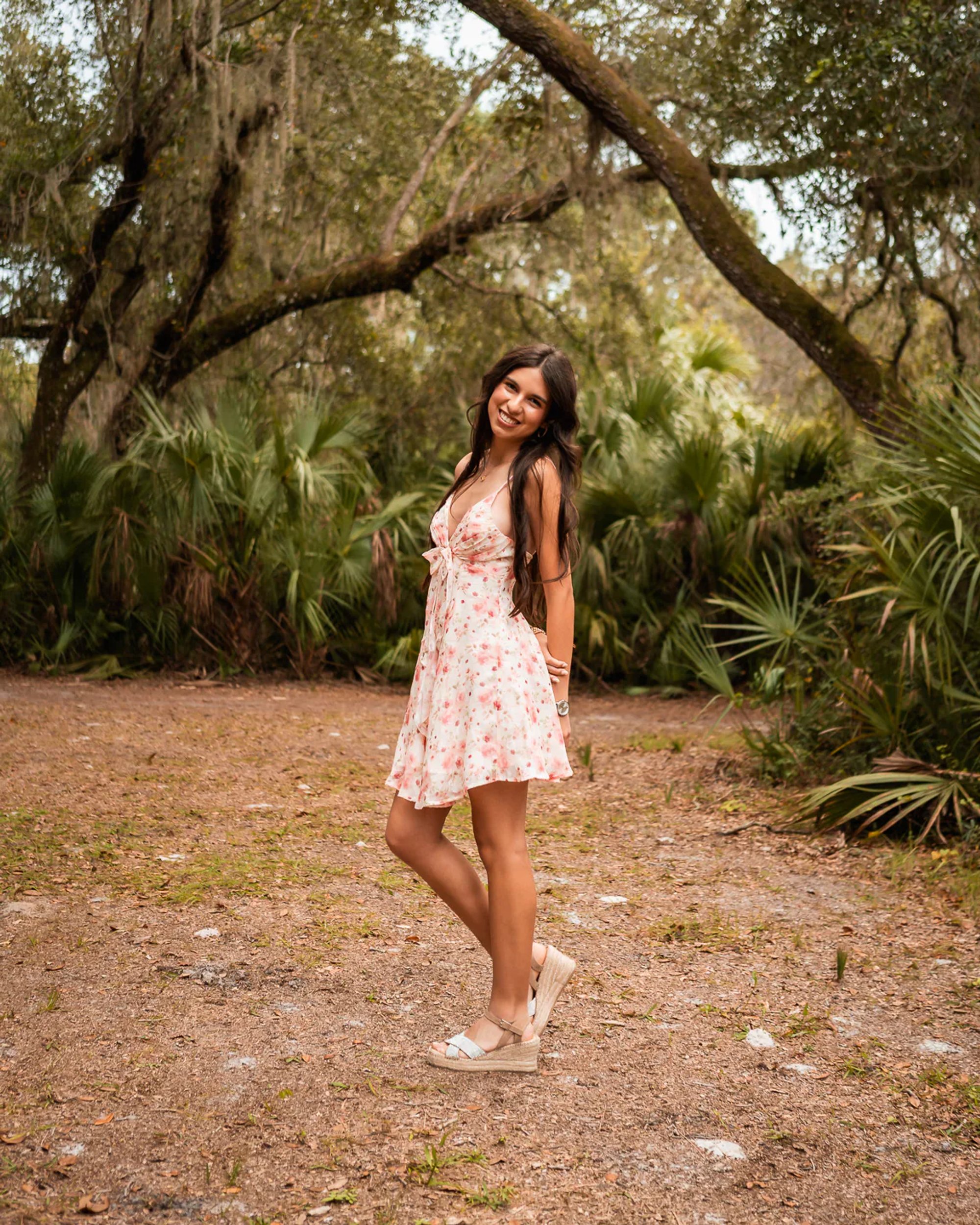 Young woman in a floral dress leaning beside a tree.
