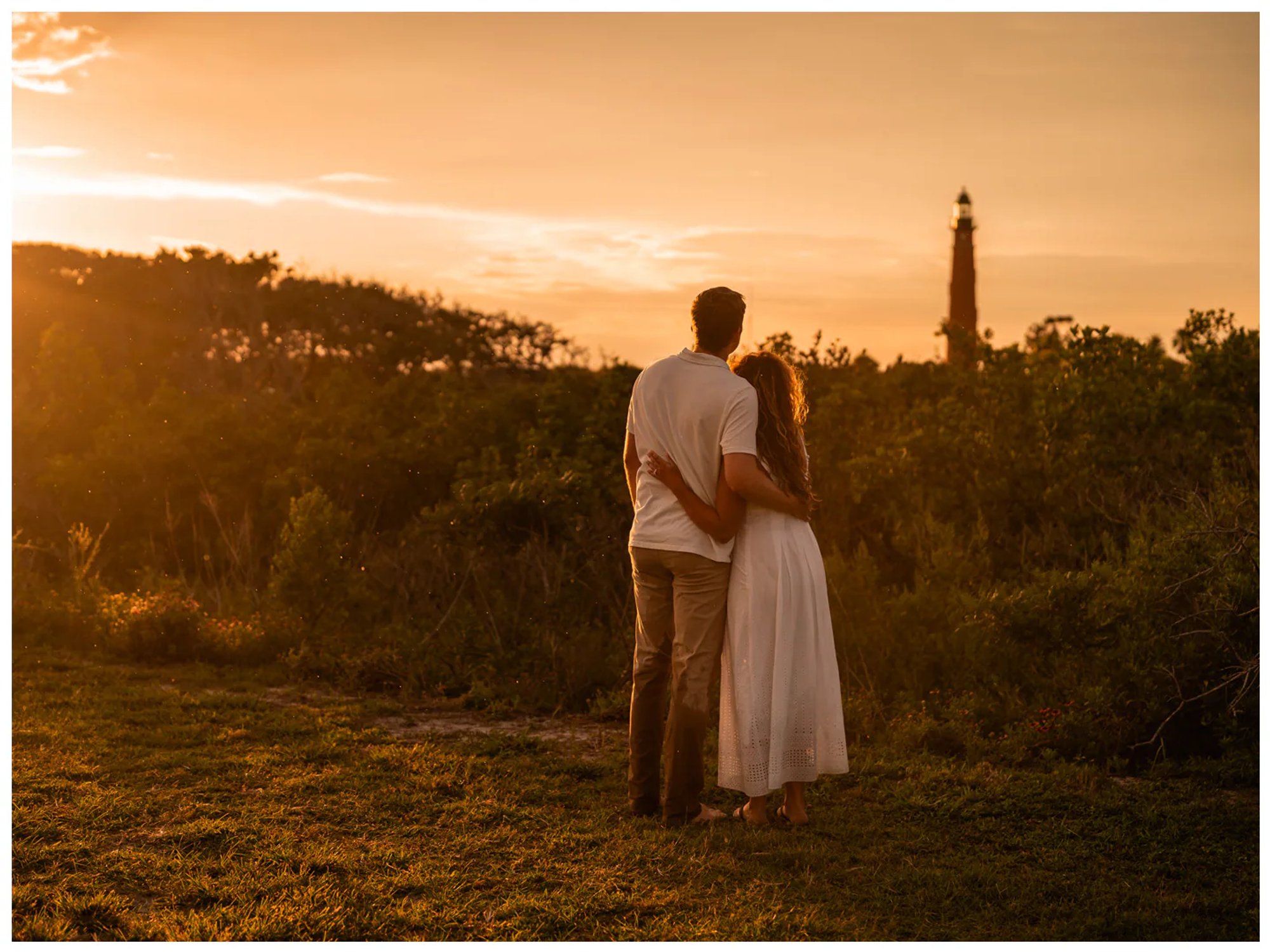 Couple embracing while looking toward a distant lighthouse at sunset.