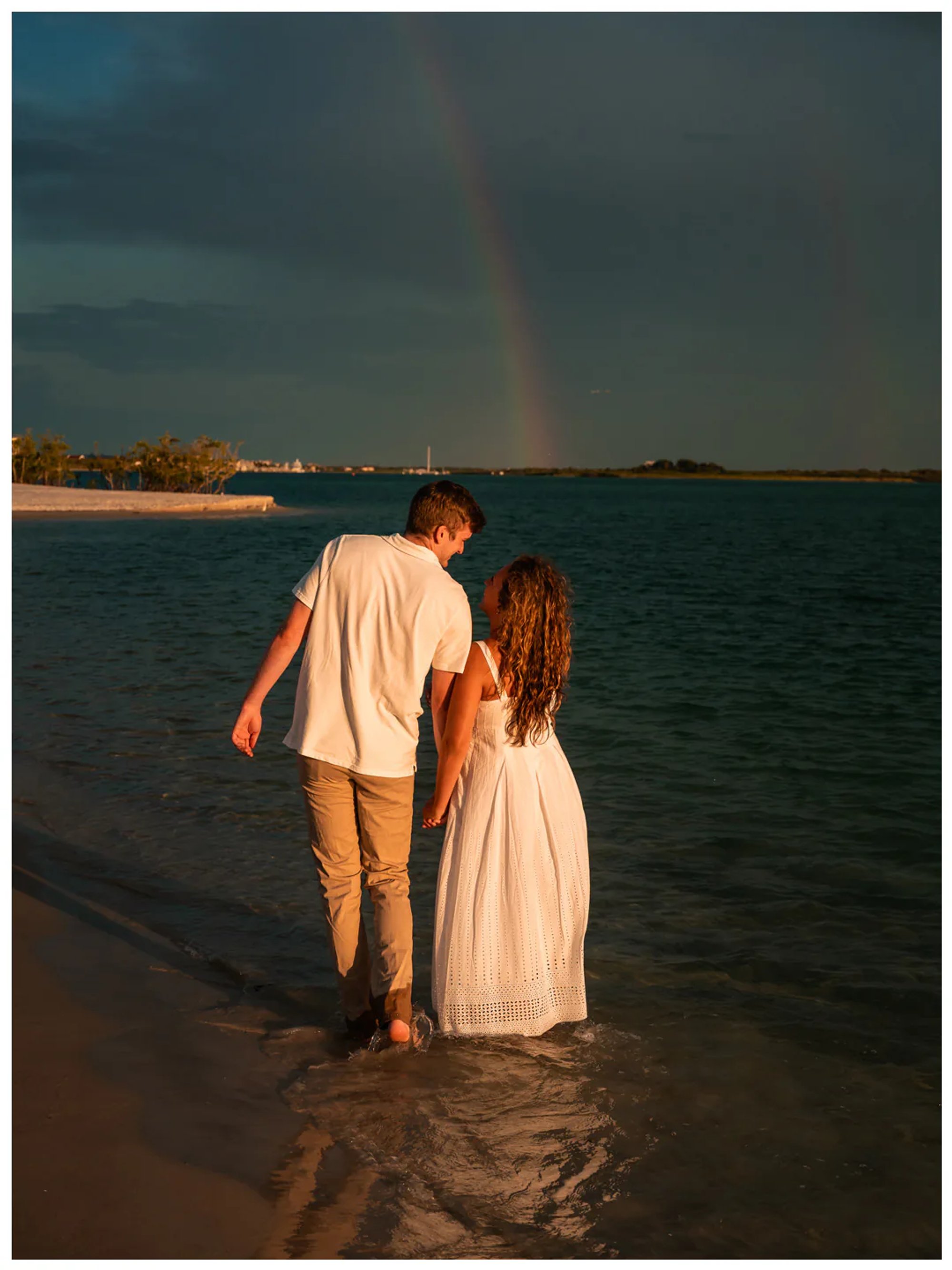 Couple walking through shallow water beneath a distant rainbow.