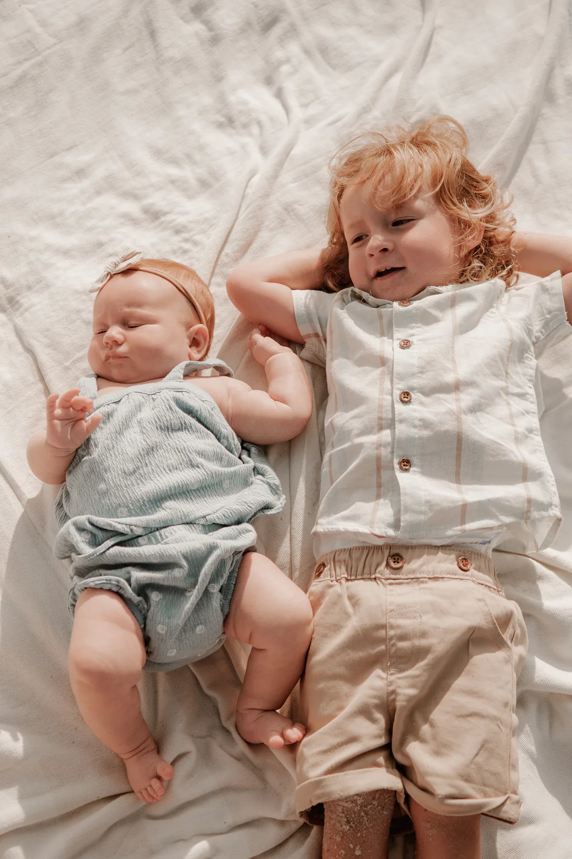 Baby and young child lying side by side on a white blanket.
