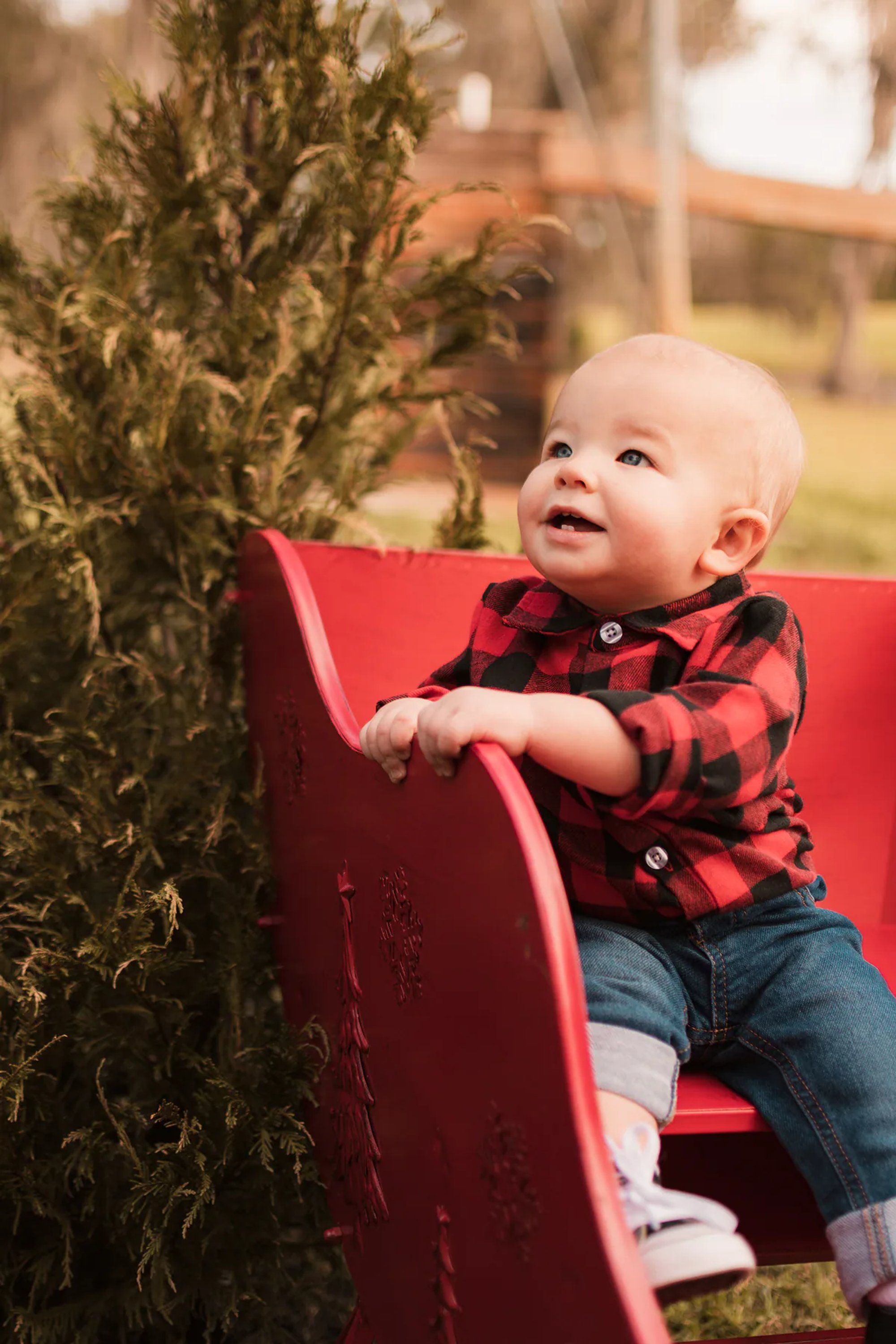Baby in red-and-black plaid seated in a small red chair outdoors.