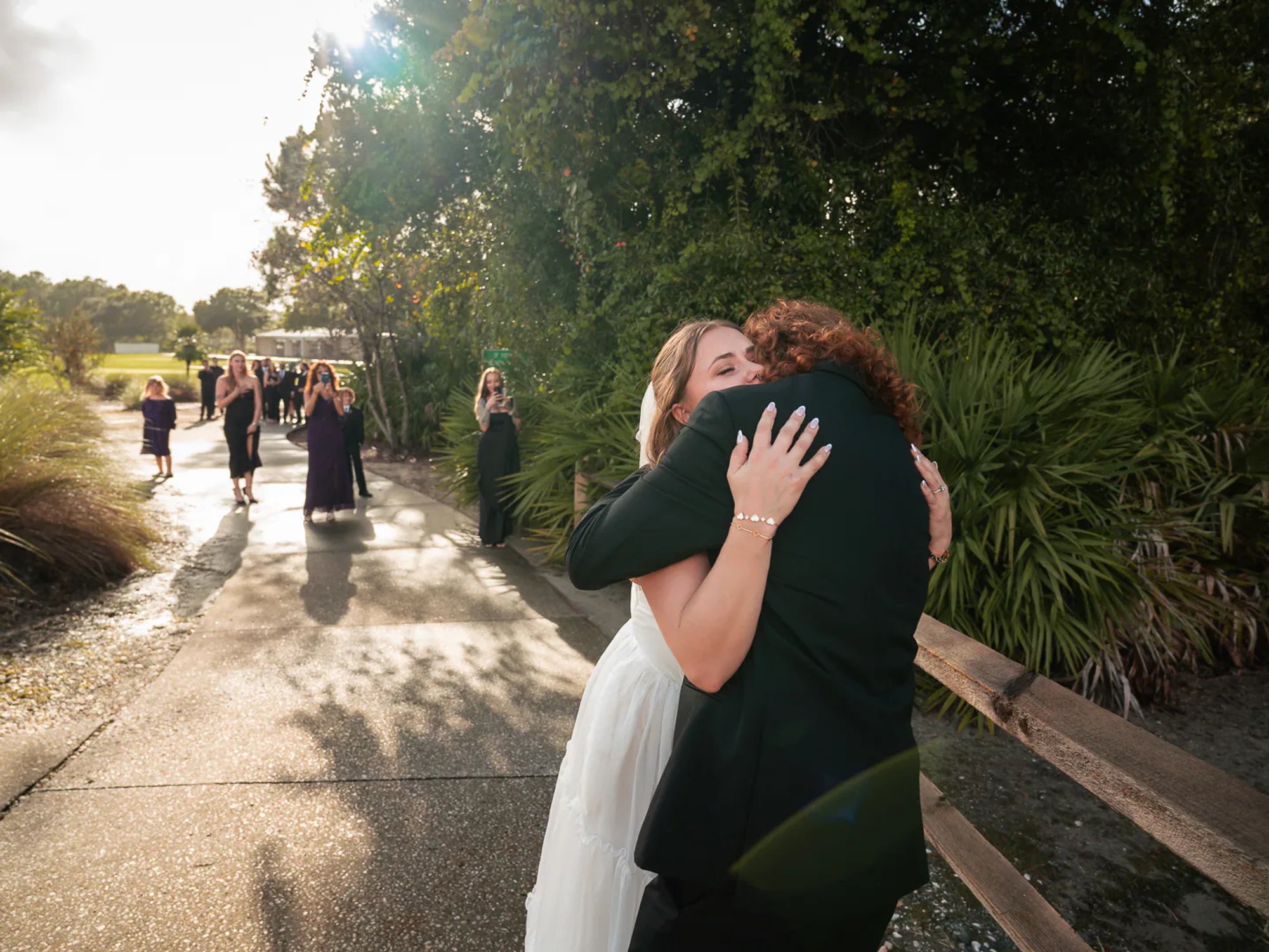 Bride embracing a wedding guest on a sunlit outdoor path.