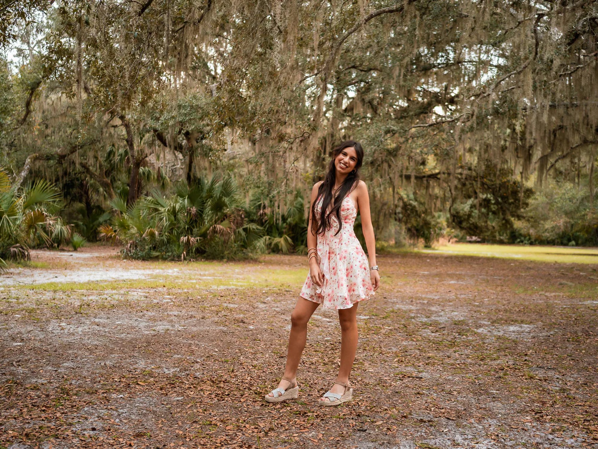 Young woman in a floral dress posing in an open wooded area.