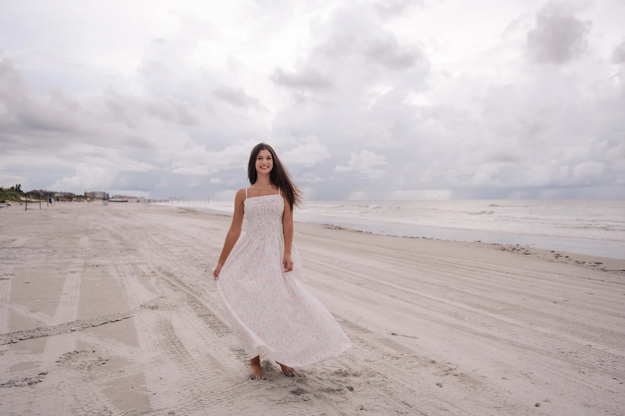 Daytona Beach senior portraits, Mia walking along the shoreline in a flowy white floral dress with overcast skies.