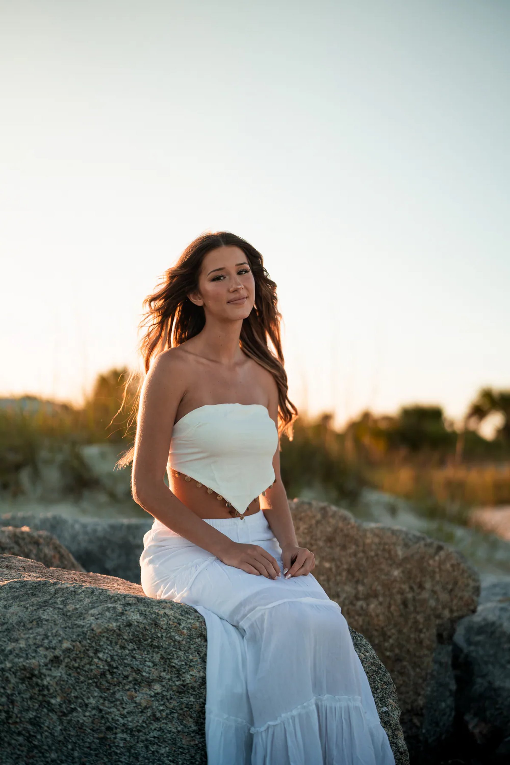 Young woman in a white dress seated on a large shoreline rock.