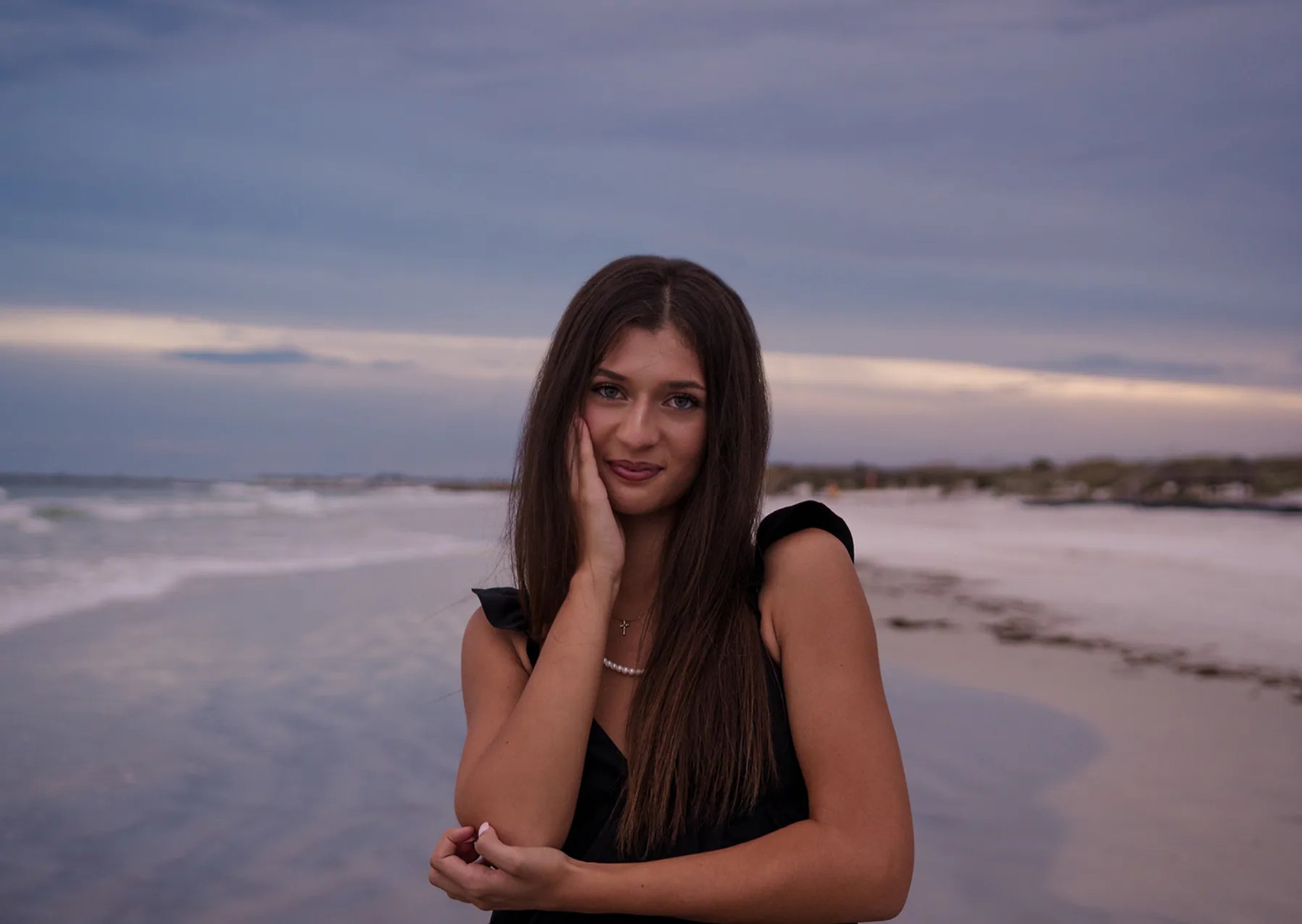 Close-up senior portrait at Ponce Inlet Beach, Mia in a black dress with ocean sunset behind her.