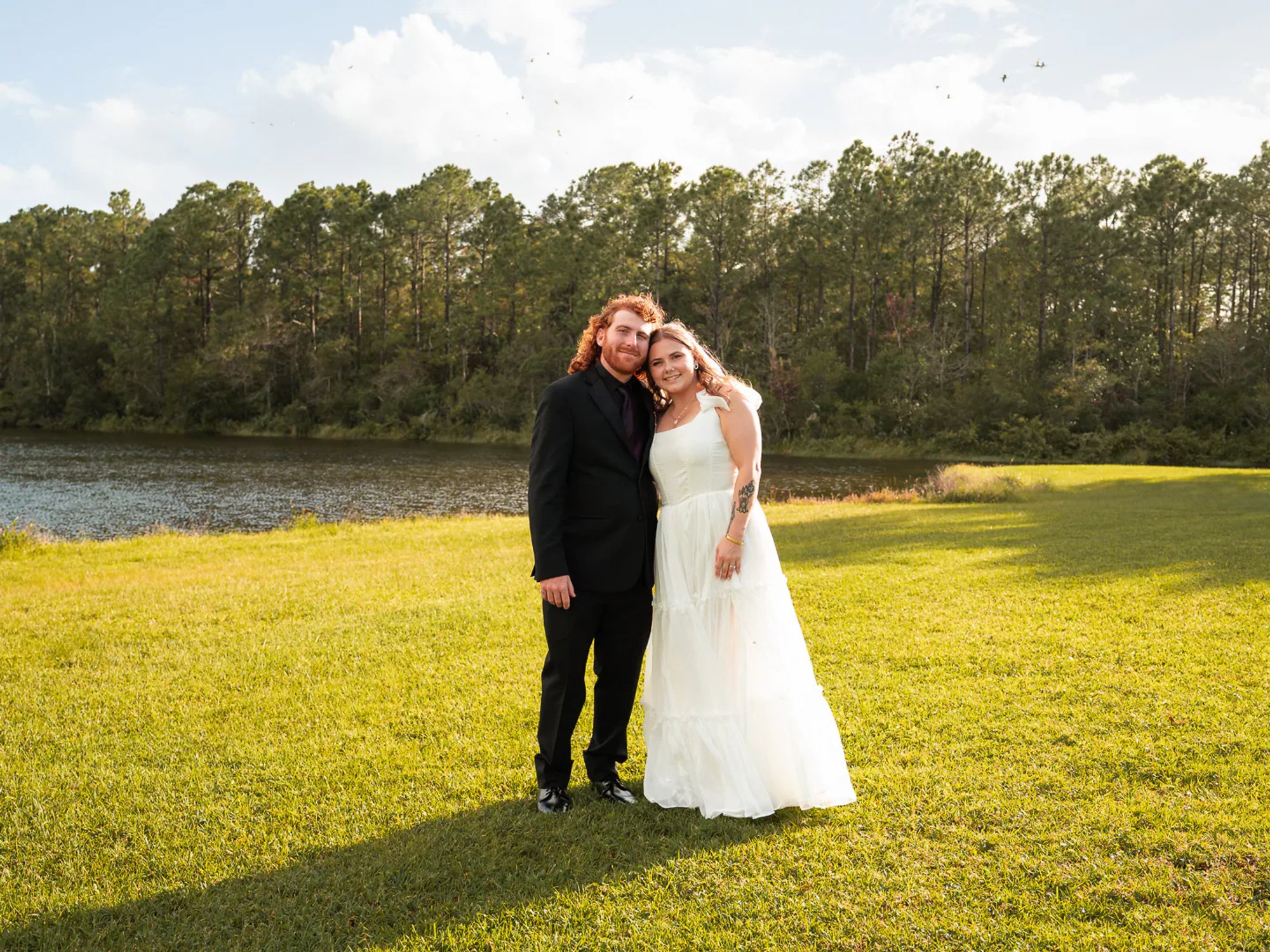 Bride and groom standing together on a sunny lawn beside a pond.