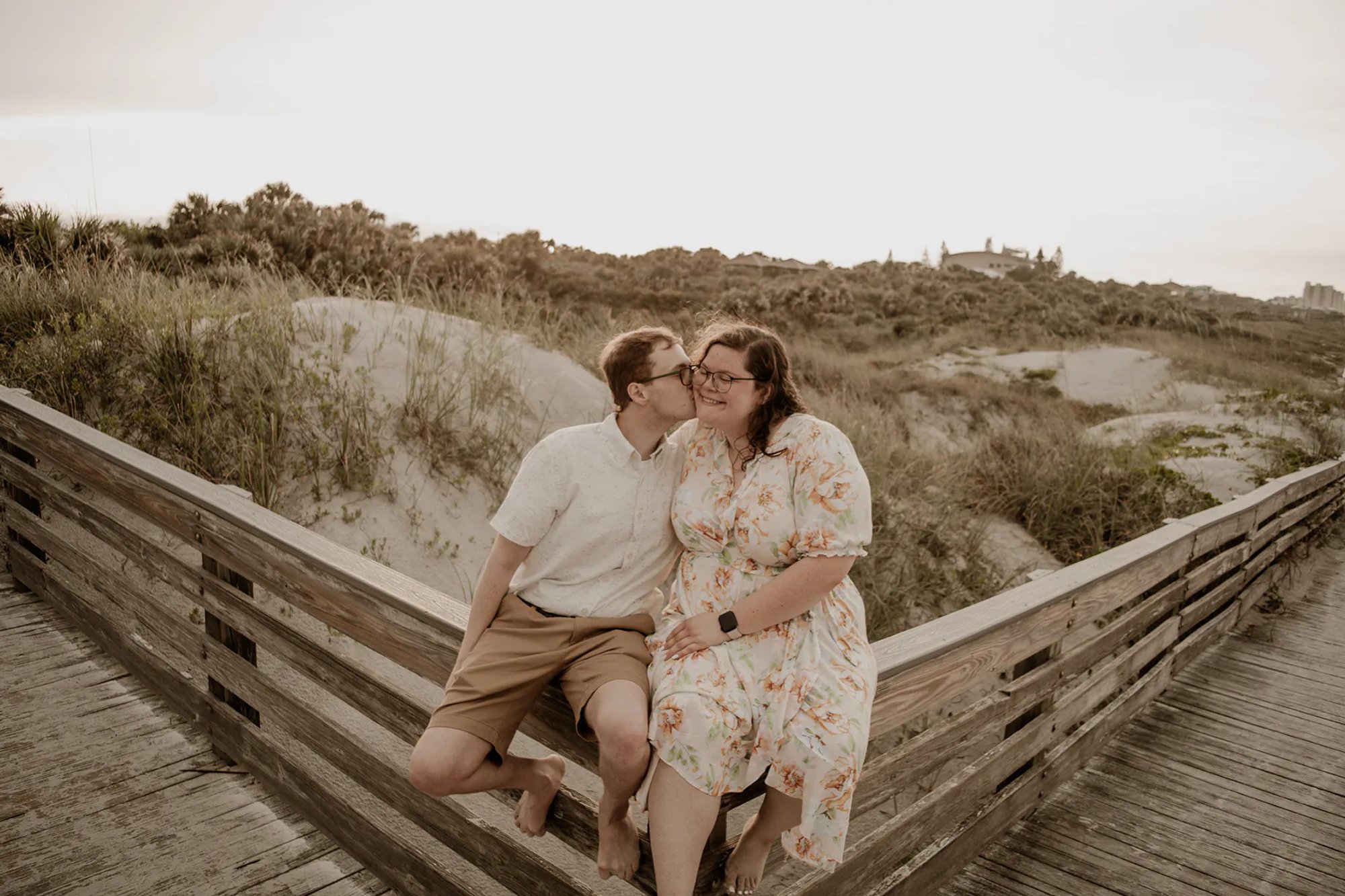 Couple sharing a kiss while seated on a wooden beach walkway.