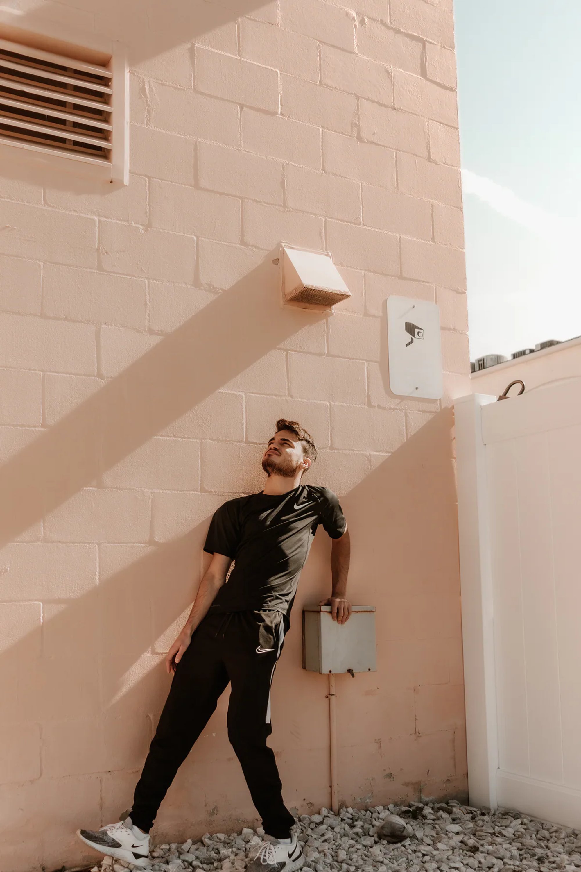Man in black clothing leaning against a pale pink industrial wall.