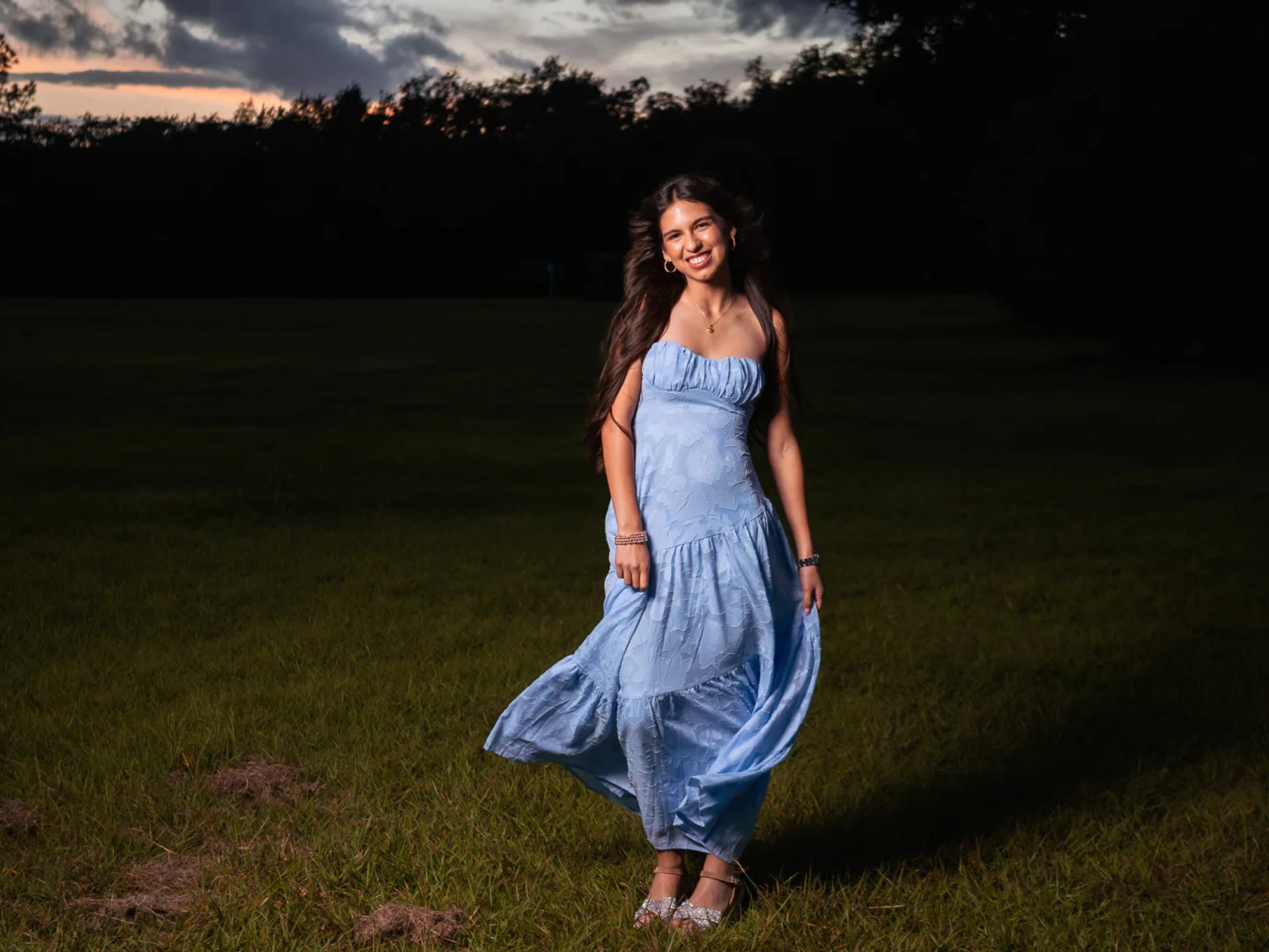 Young woman walking across a dark field in a flowing blue dress.
