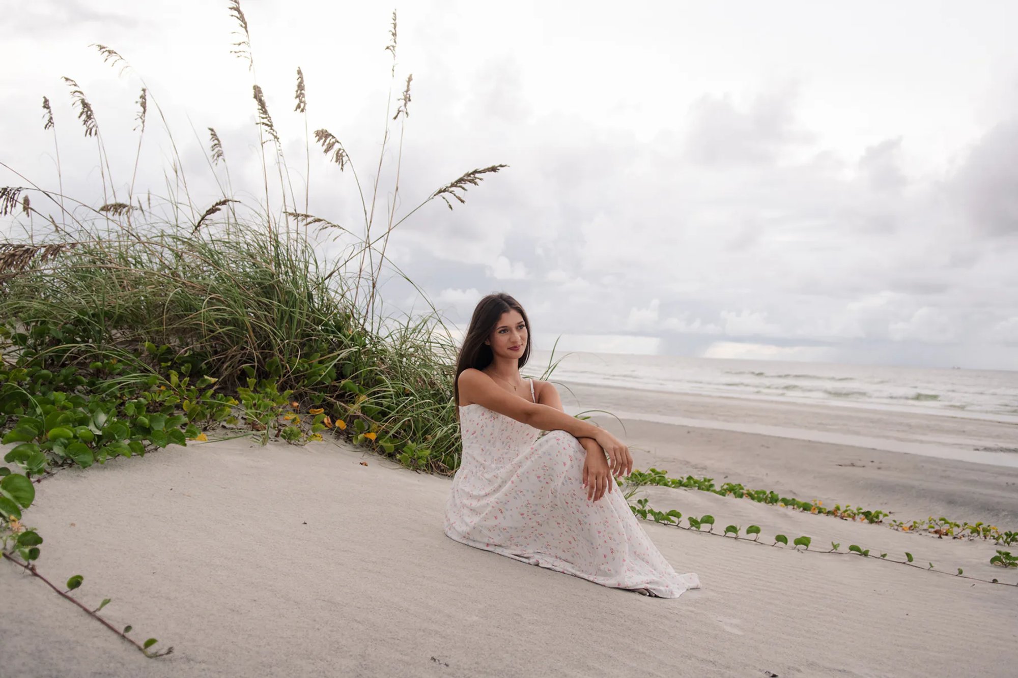 Senior photos in Ponce Inlet FL, Mia sitting in a white floral dress near the dunes with the beach in the background.