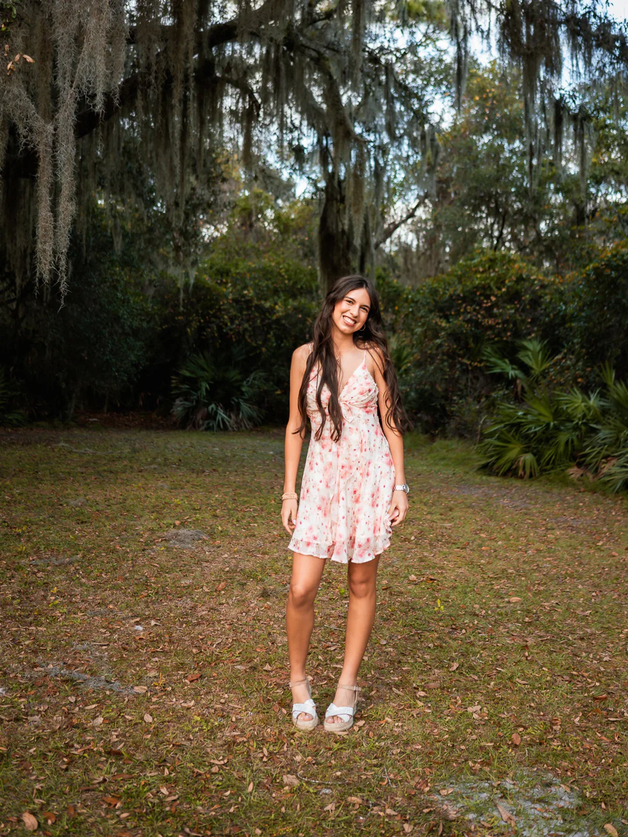 Young woman in a pink floral dress standing in a wooded clearing.