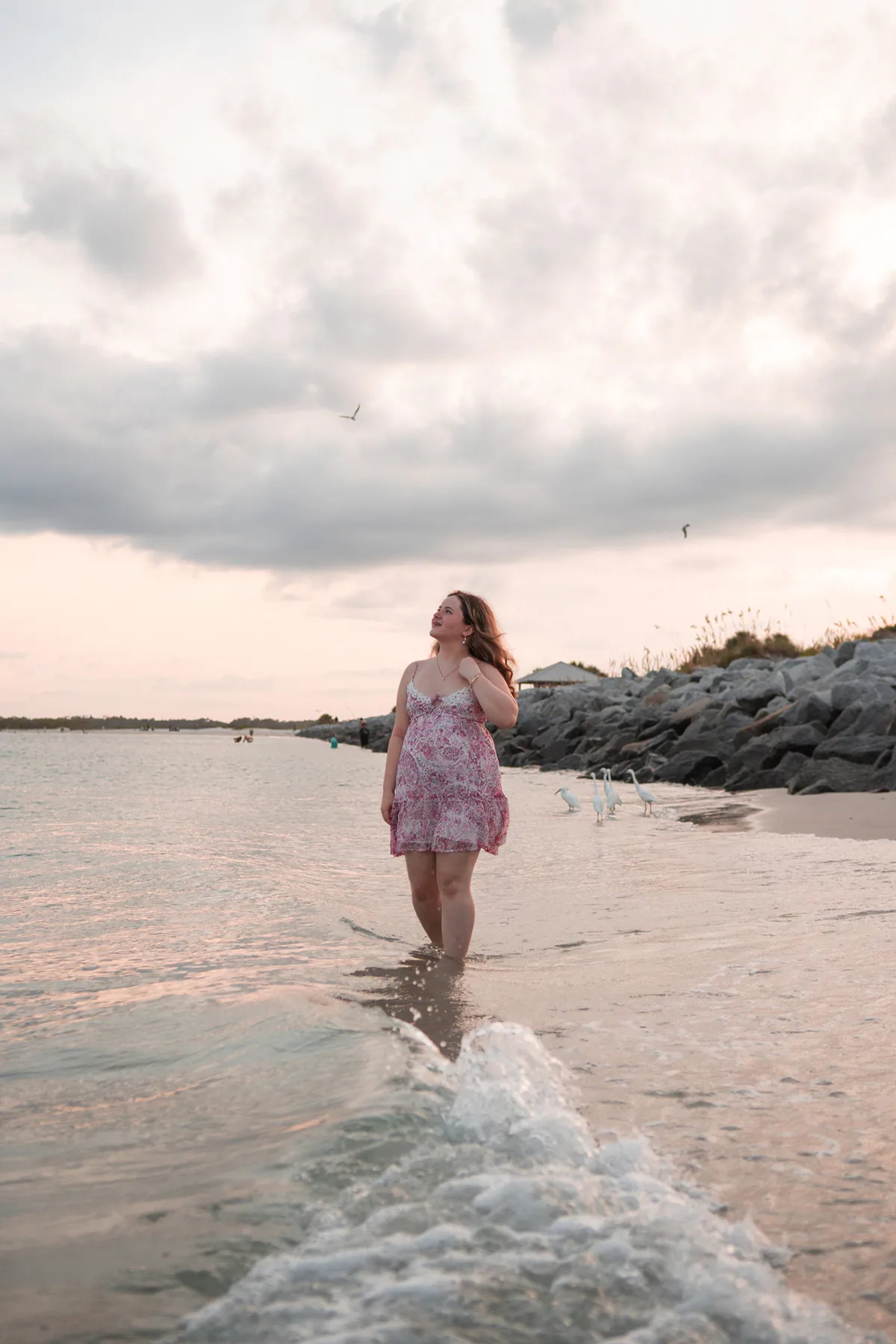 Young woman in a pink floral dress walking through the surf.