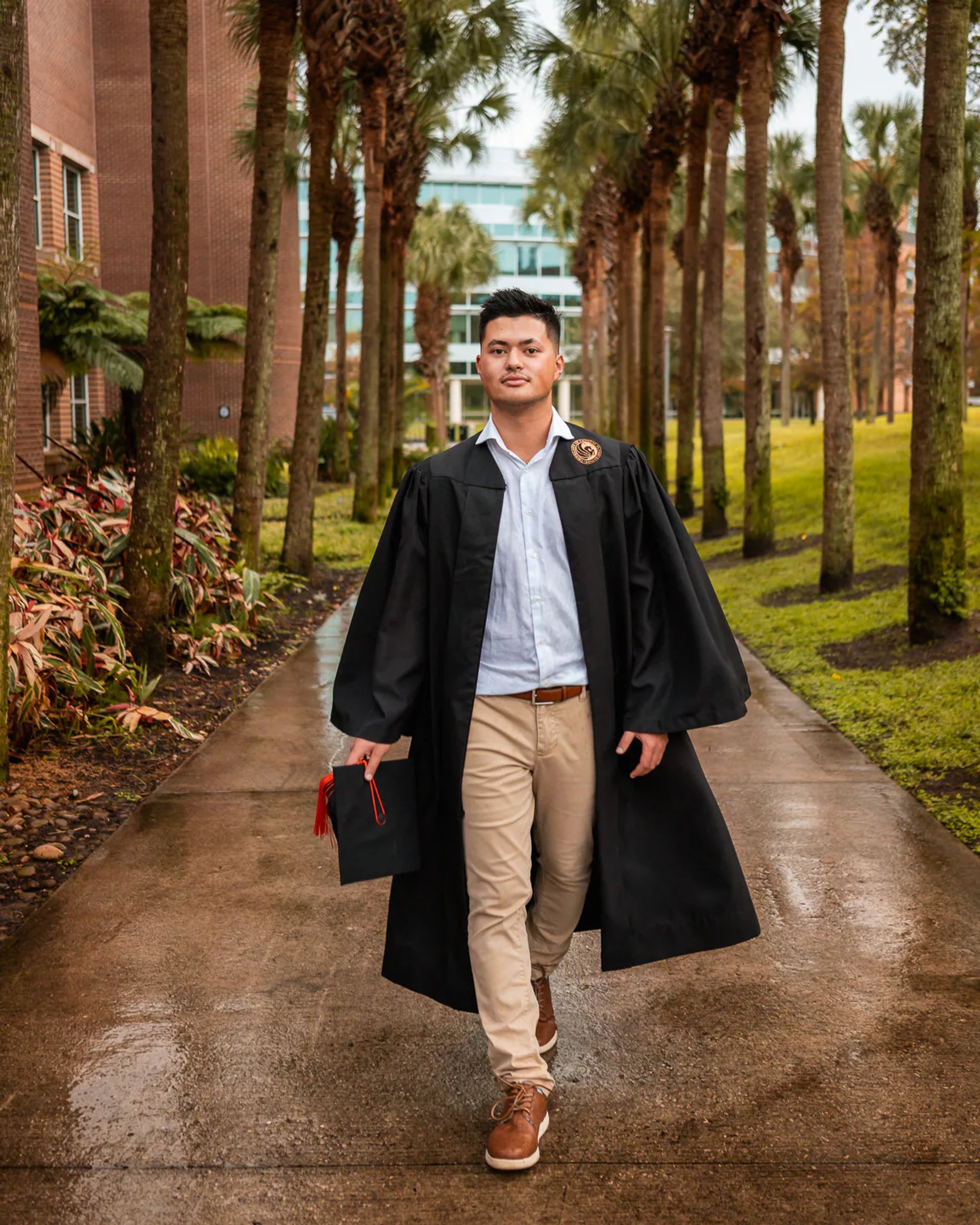 Male graduate walking along a palm-lined path in his black gown.