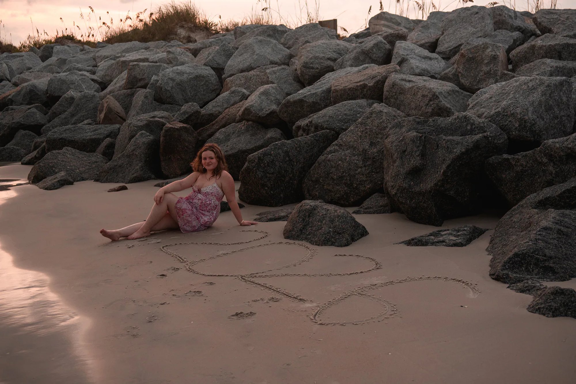 Senior photo in Ponce Inlet FL, Nelly sitting by large rocks on the shoreline, pink floral dress at golden hour.