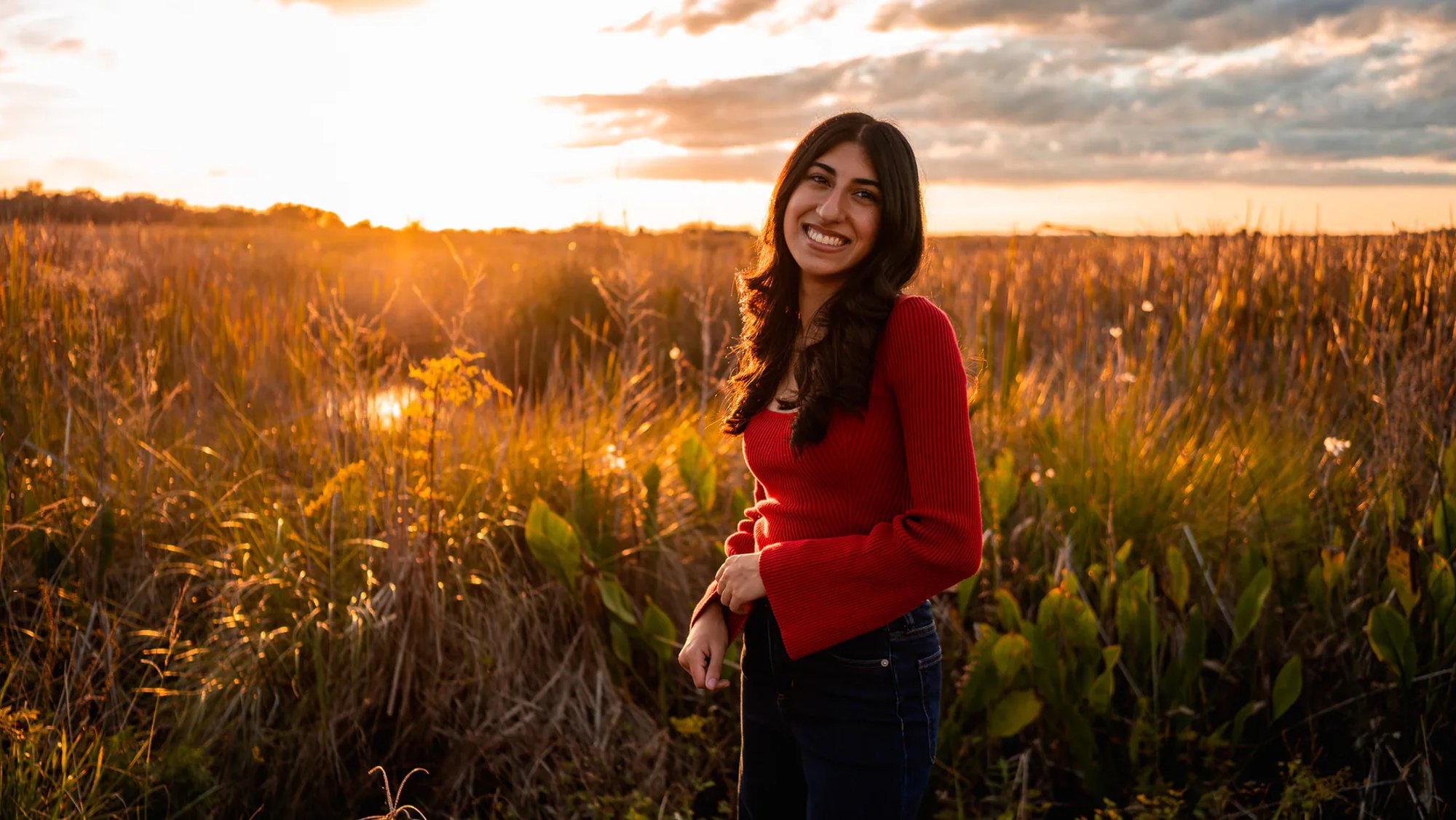 Young woman in a red top standing among marsh grass at sunset.