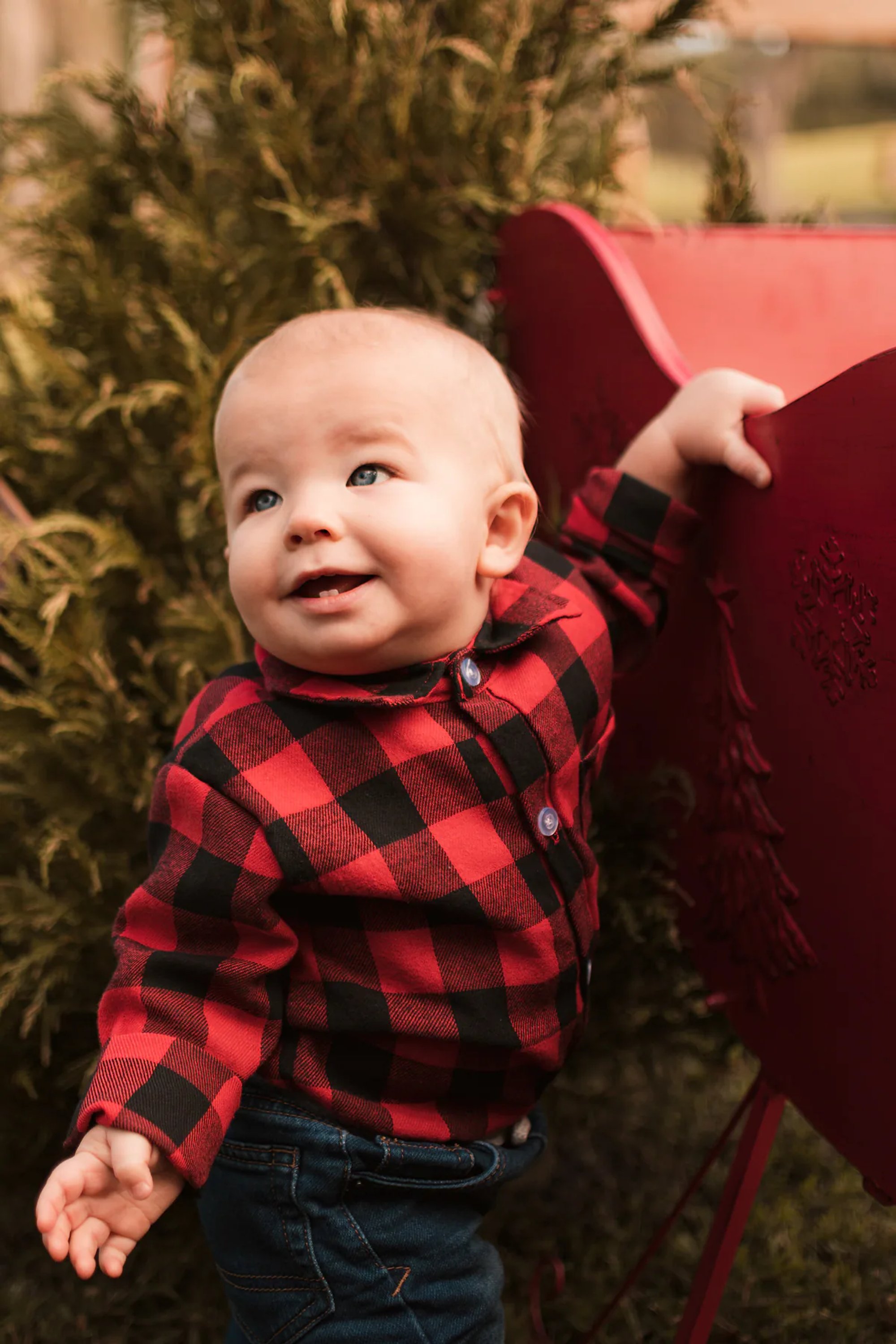 Smiling baby in red-and-black plaid beside a red chair.