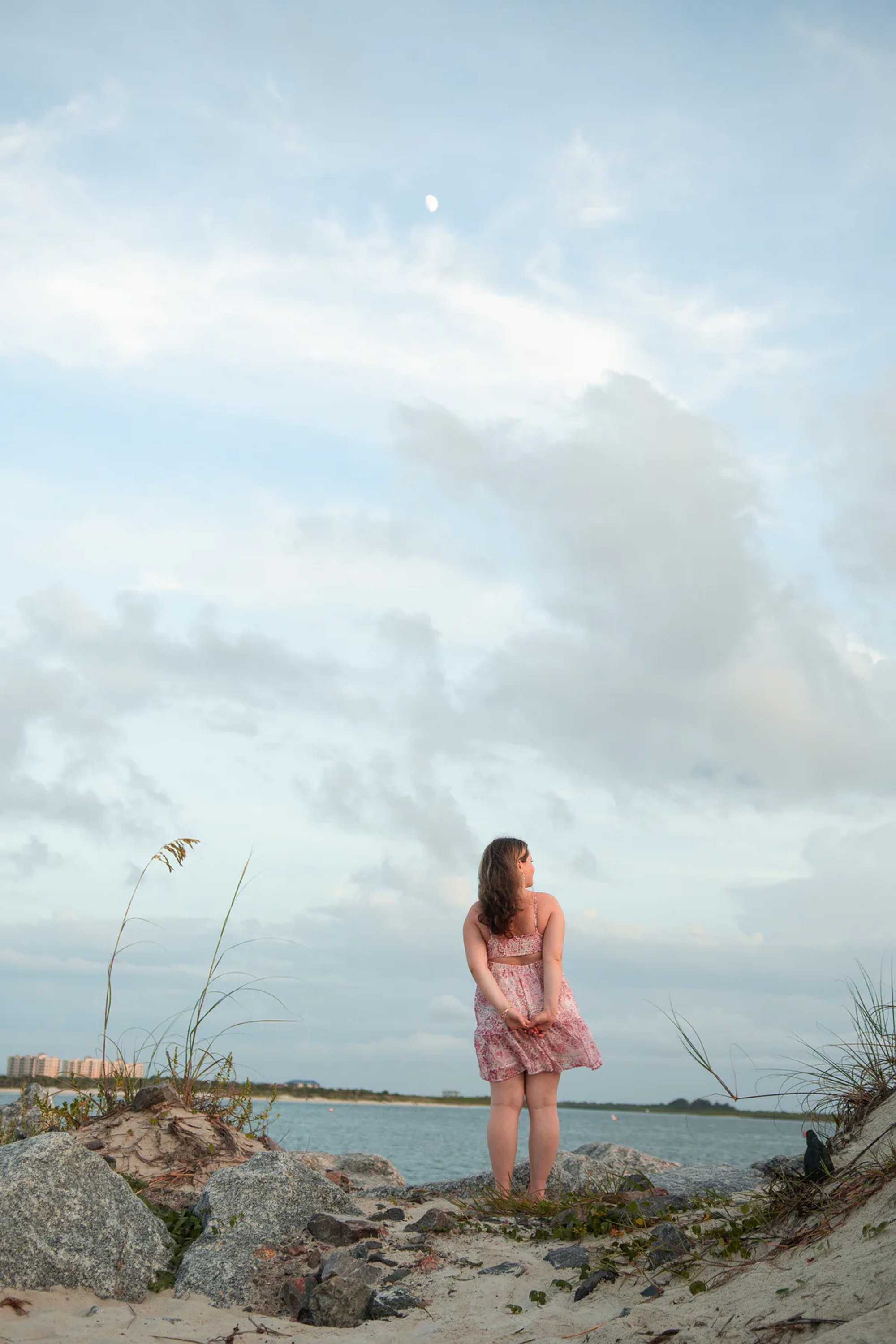 Young woman in a pink floral dress standing on shoreline rocks.
