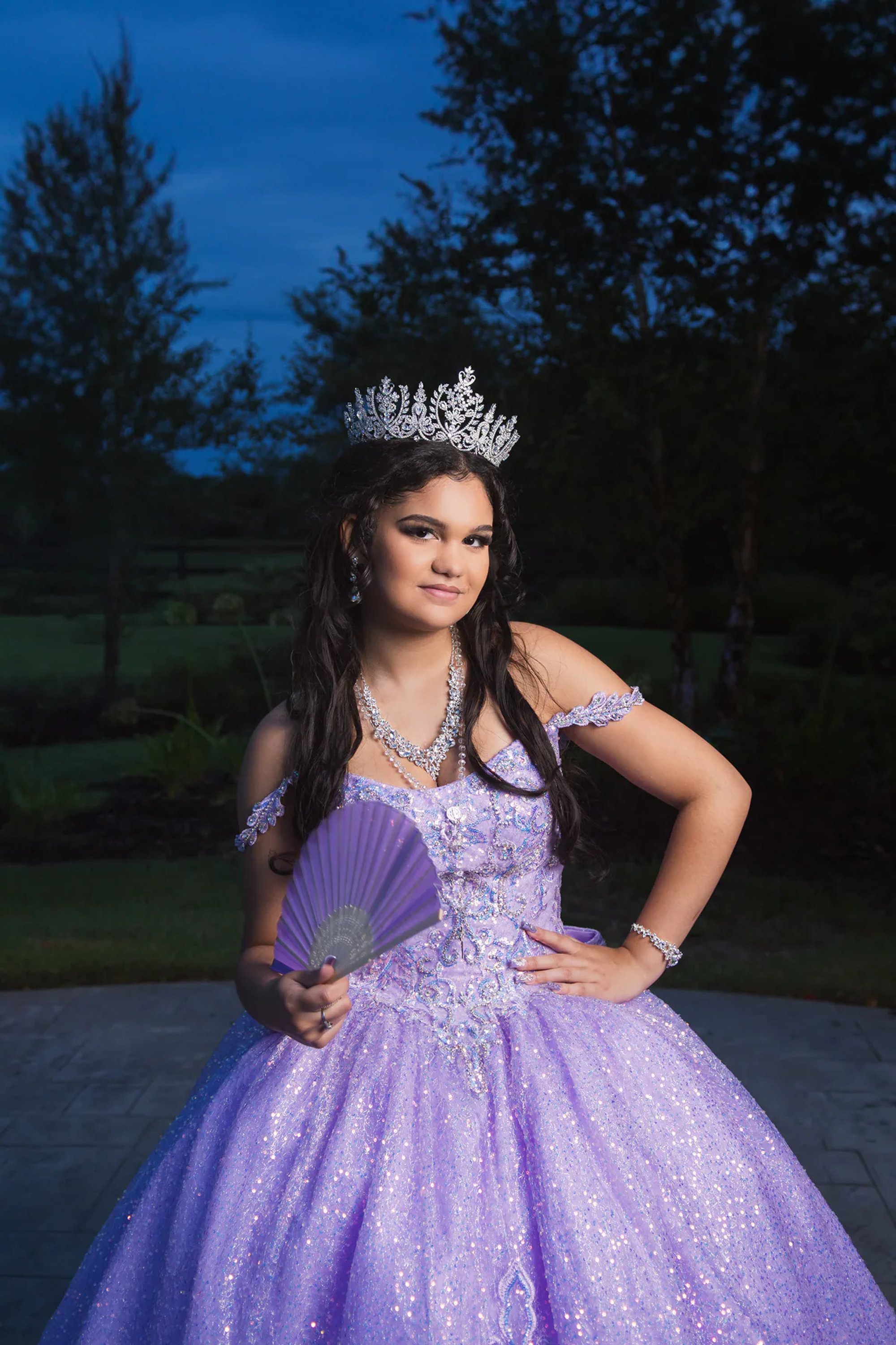 Quinceañera posing outdoors with a purple fan and sparkling tiara.