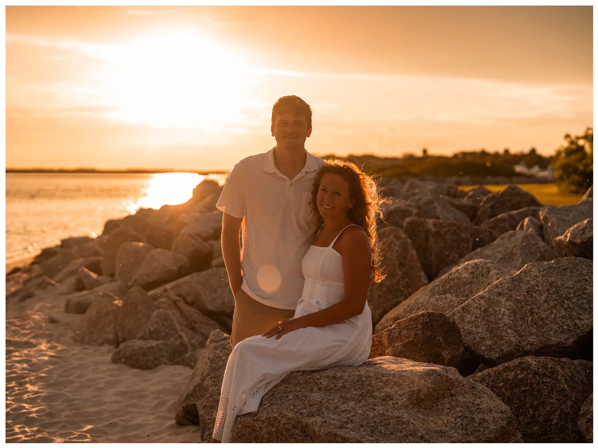 Couple seated together on large shoreline rocks at sunset.