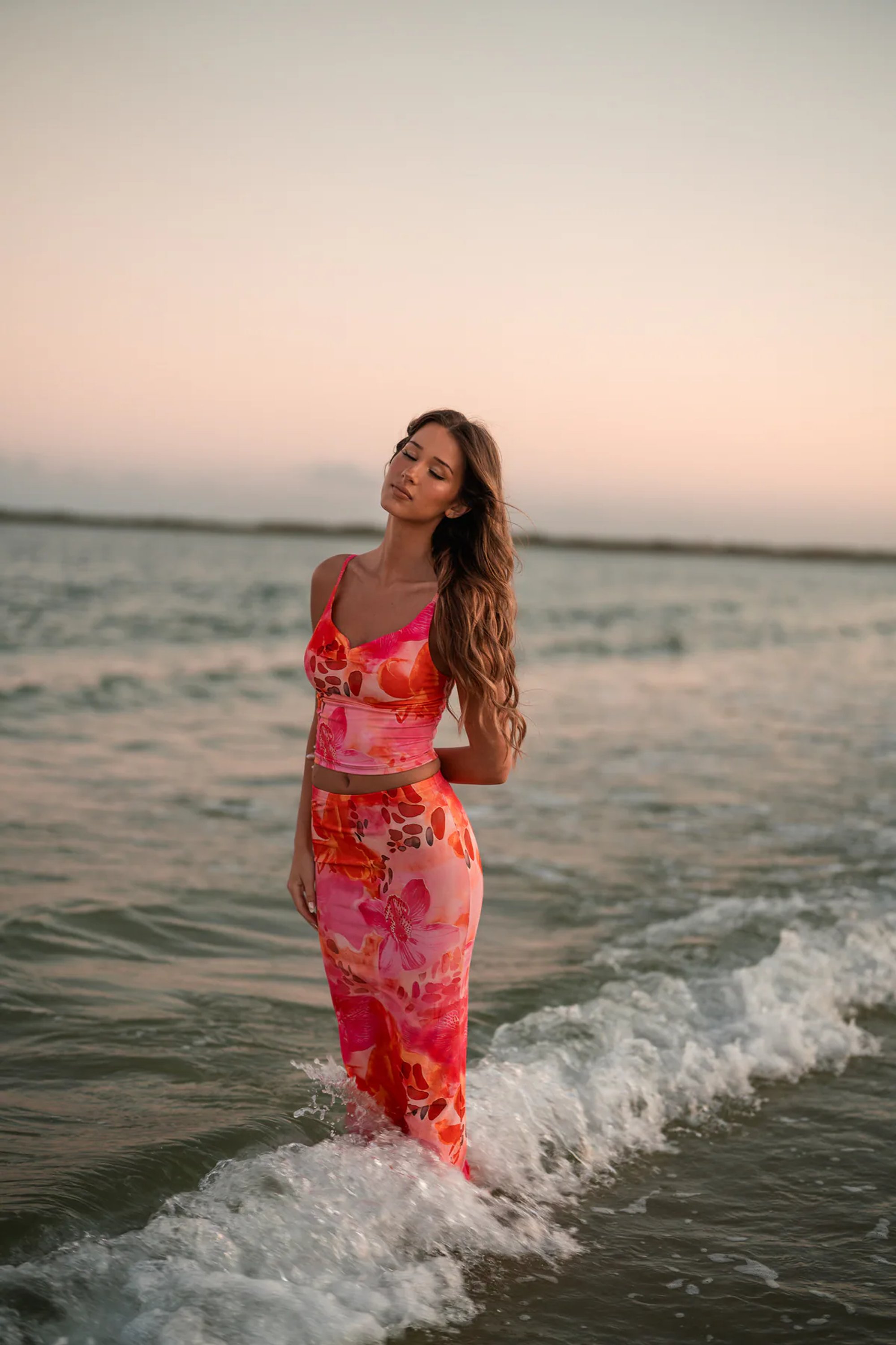 Young woman in a coral patterned dress standing in the ocean surf.