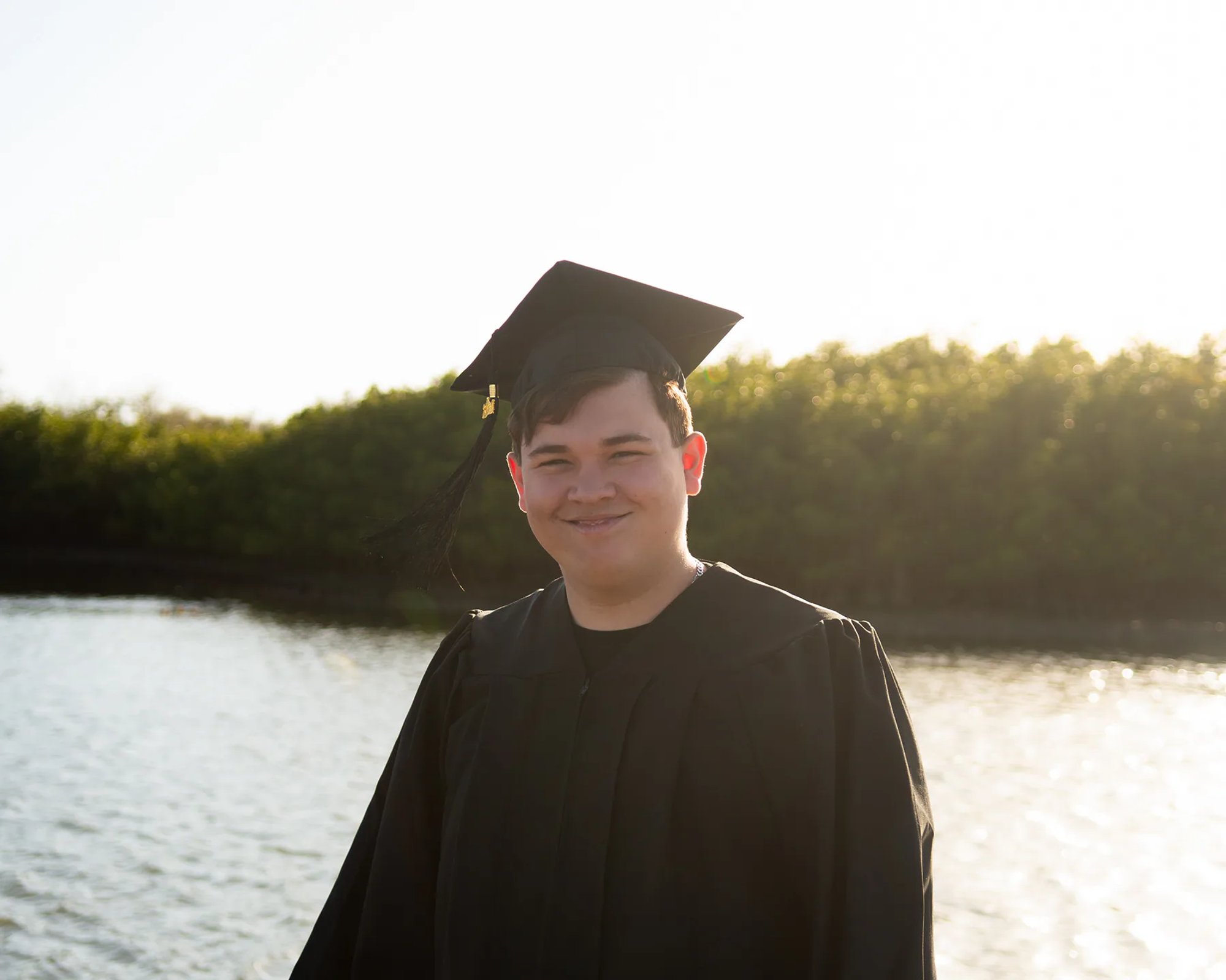 Graduate in a black cap and gown smiling beside a sunlit lake.