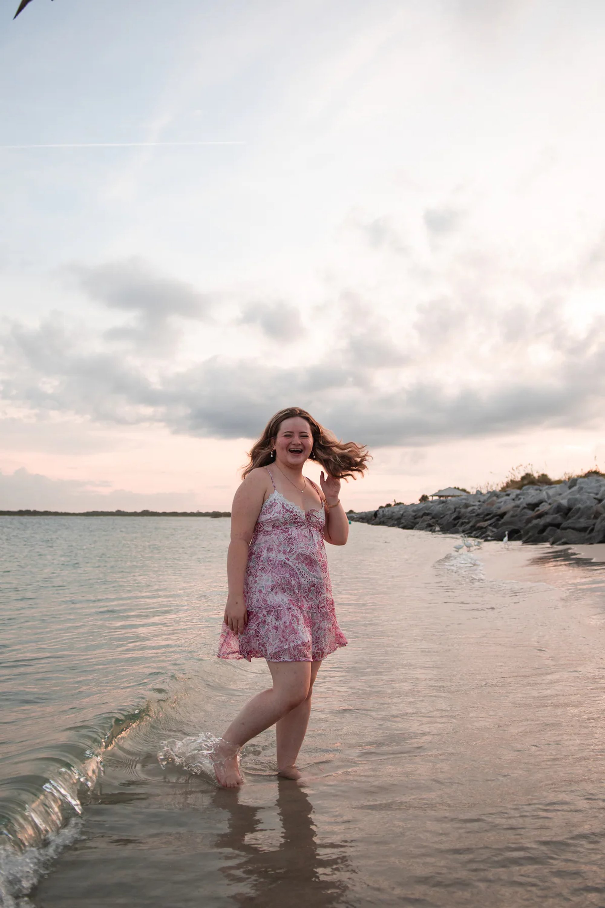 Senior portraits at Daytona Beach area, Nelly walking along shoreline rocks in pink dress with pastel sunset sky.