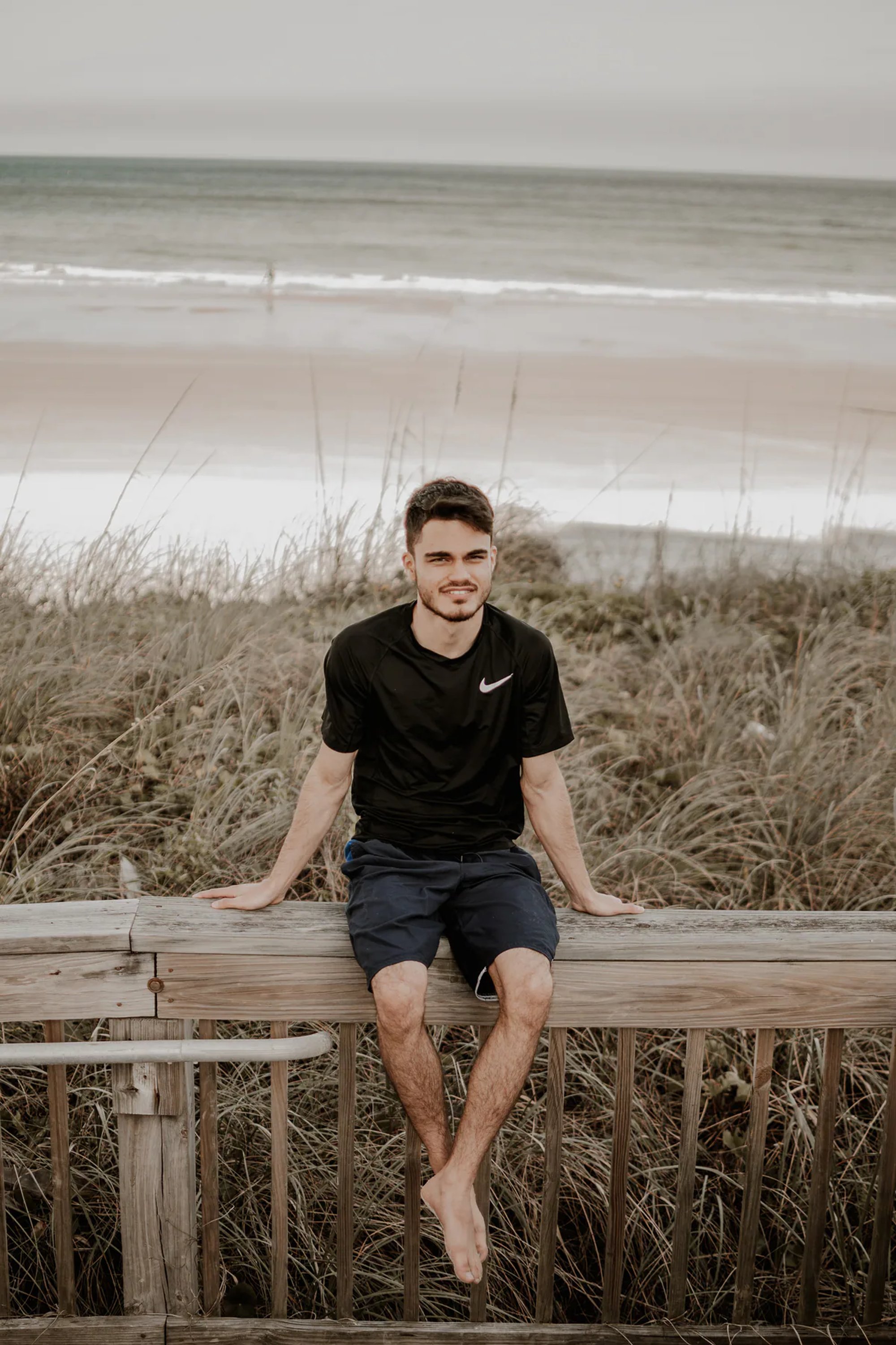 Young man seated on a boardwalk railing with the ocean behind him.