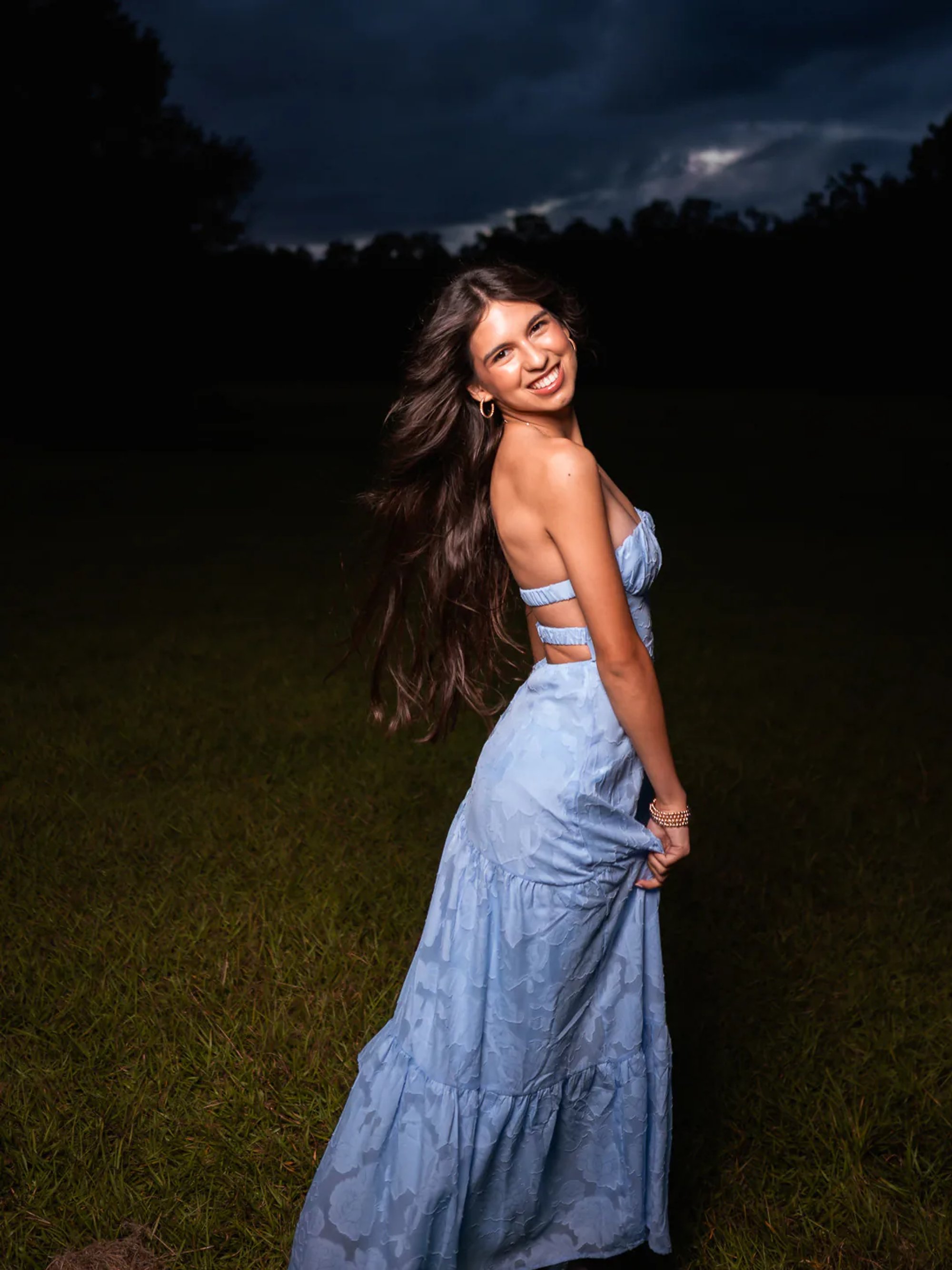 Young woman looking over her shoulder in a blue dress beneath a dramatic sky.