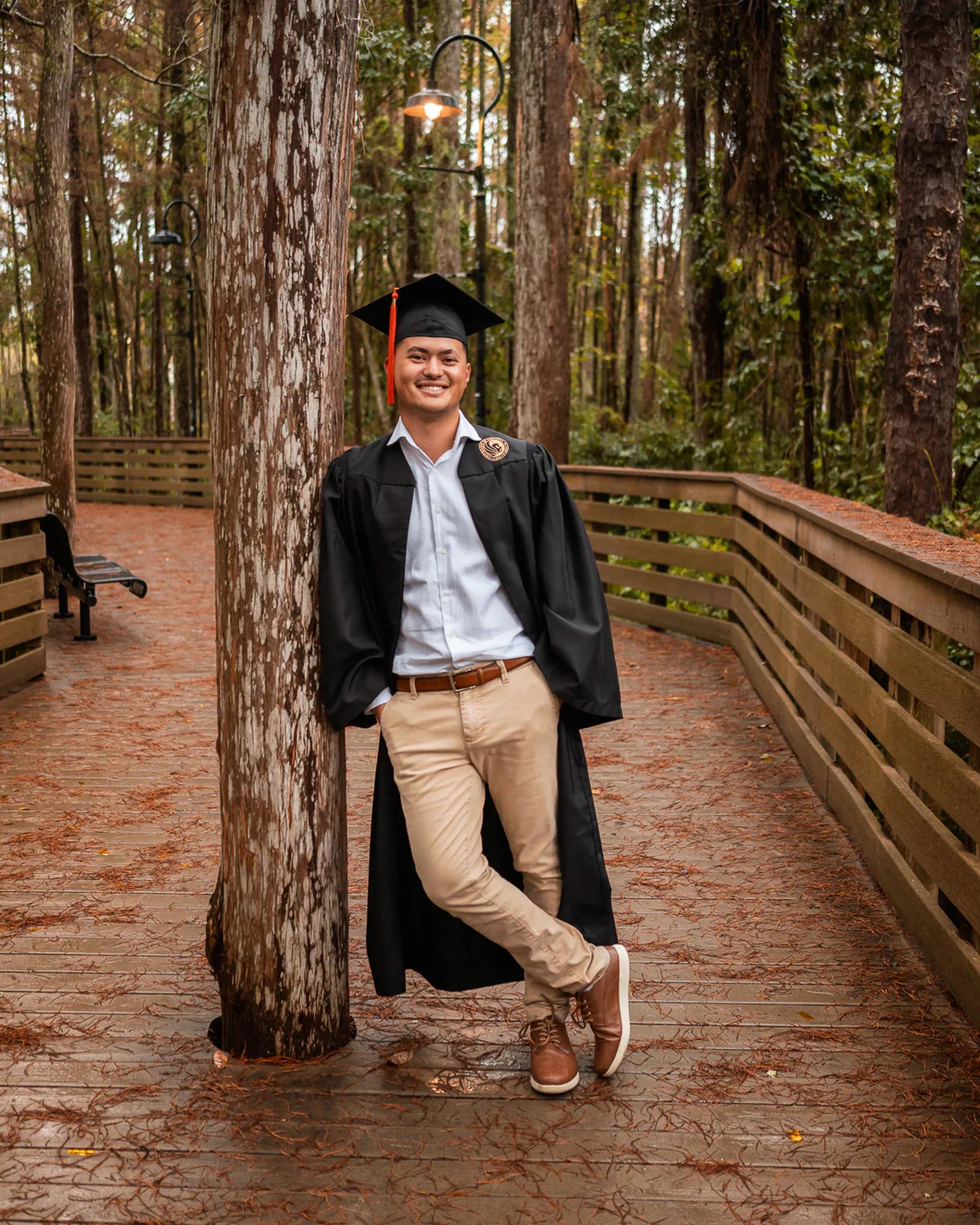 Male graduate in a black cap and gown leaning against a tree.