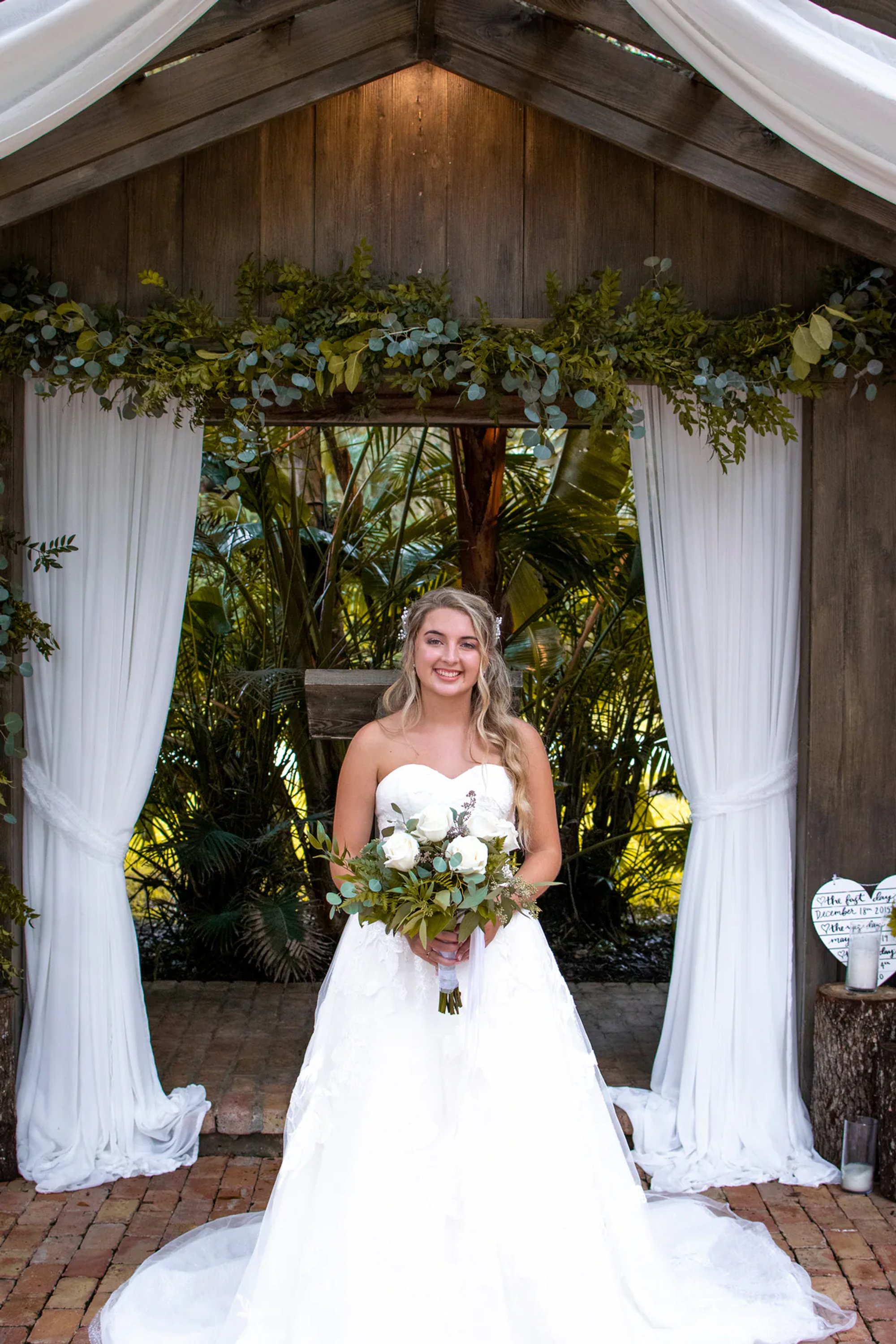 Bride standing beneath a rustic wedding arch holding a white bouquet.