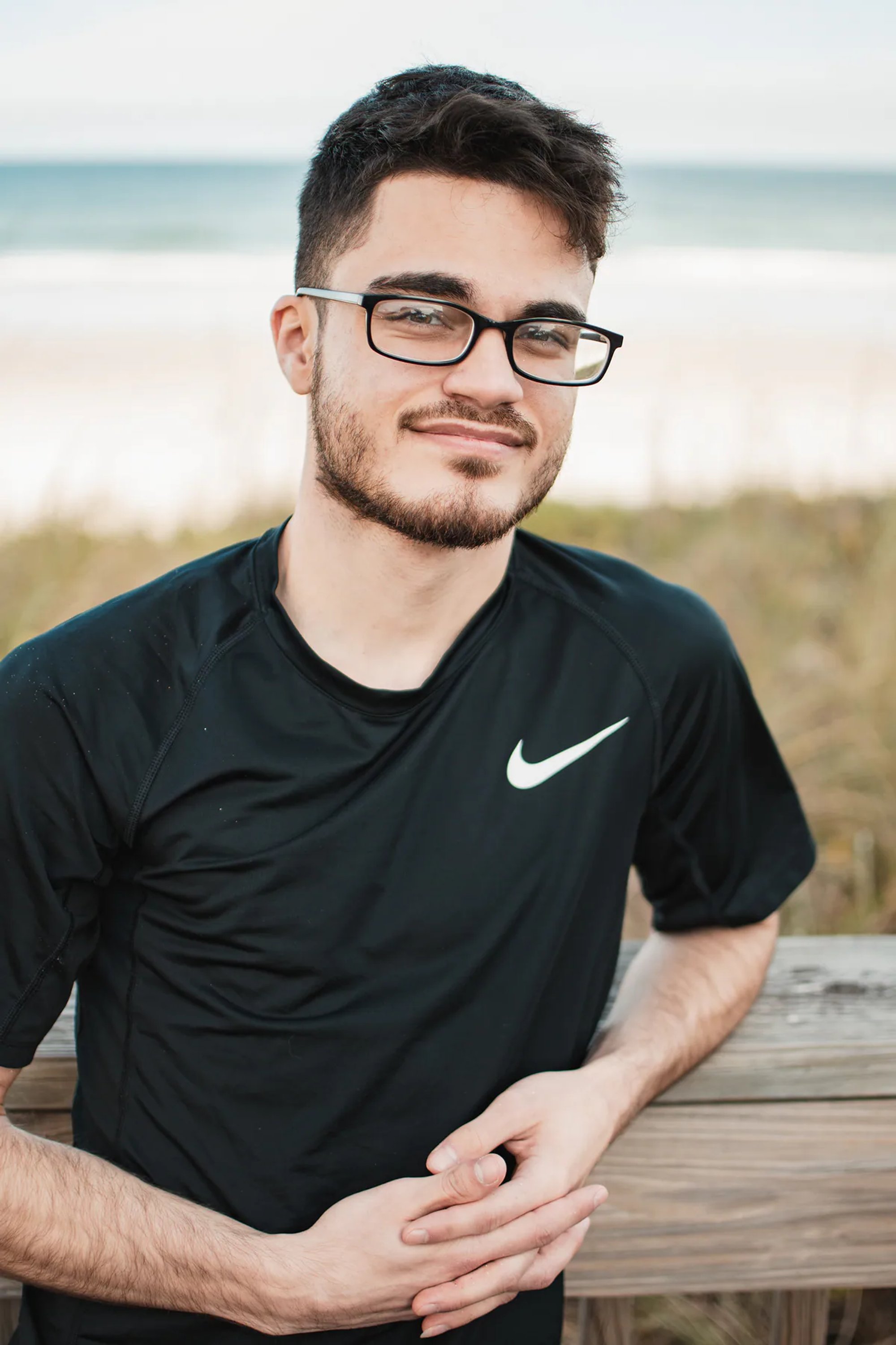 Young man with glasses leaning on a beach boardwalk railing.