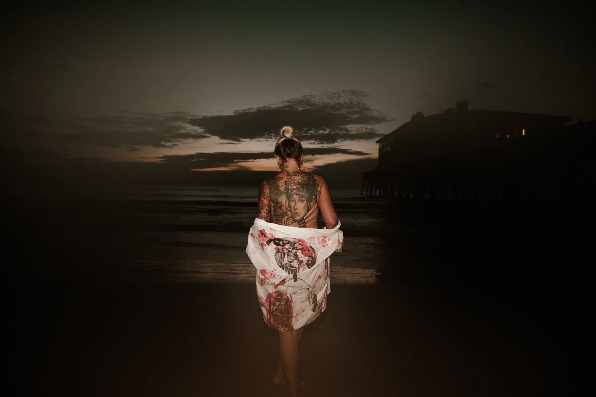 Tattooed woman holding a floral wrap while facing the ocean at dusk.