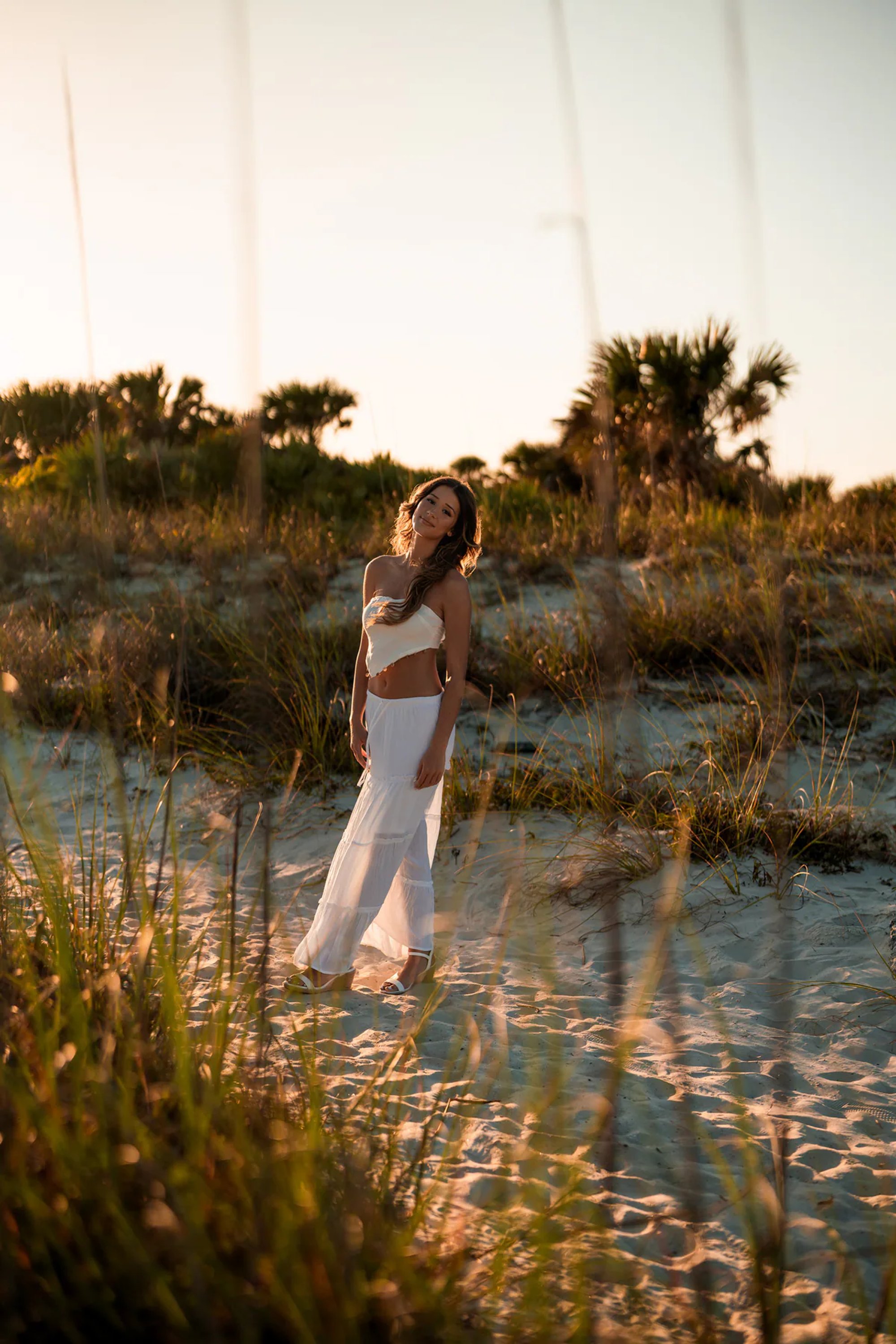 Young woman in a white outfit standing in dune grass at sunset.