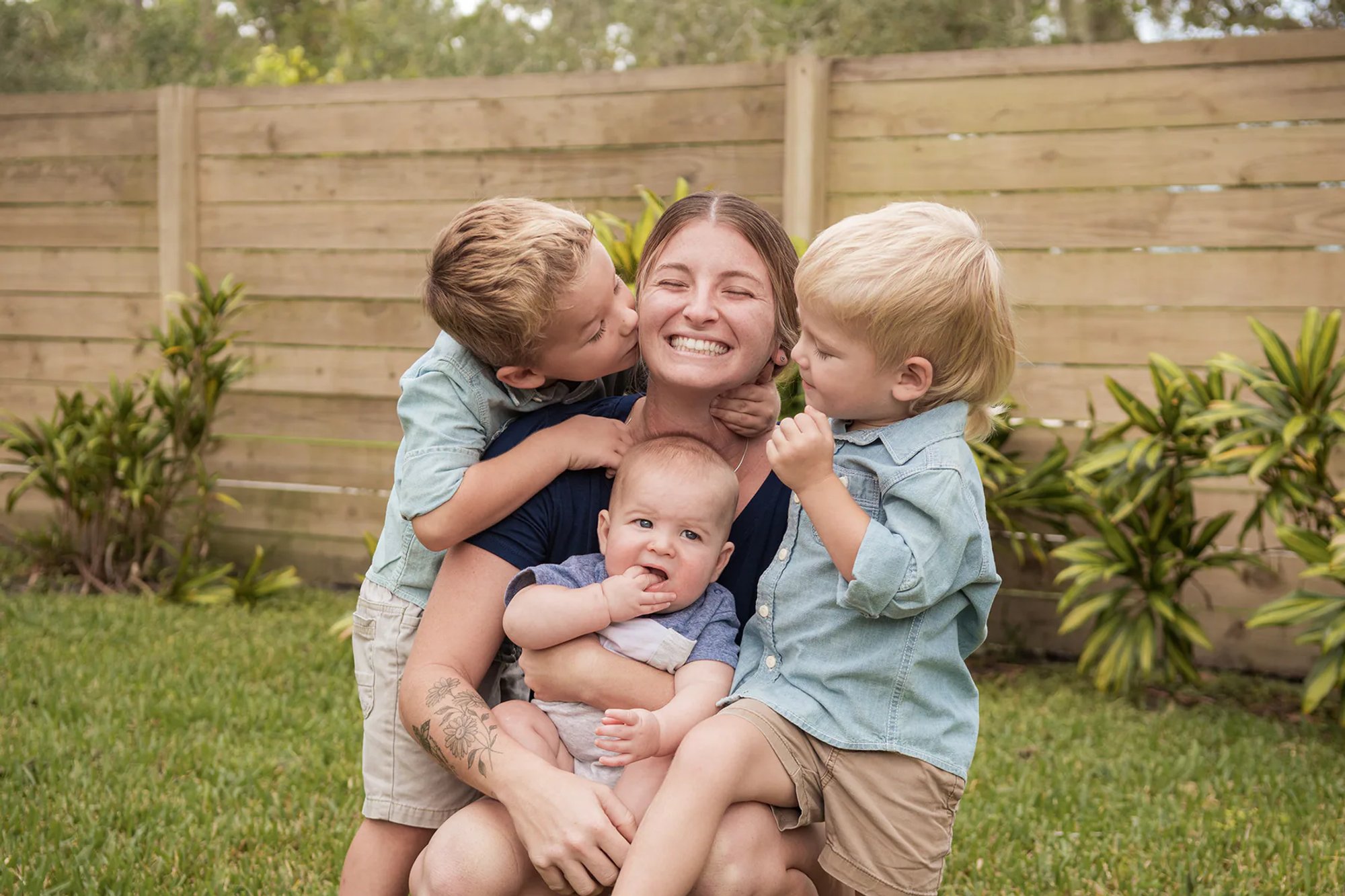 Mother laughing as two young boys kiss her cheeks while she holds a baby.