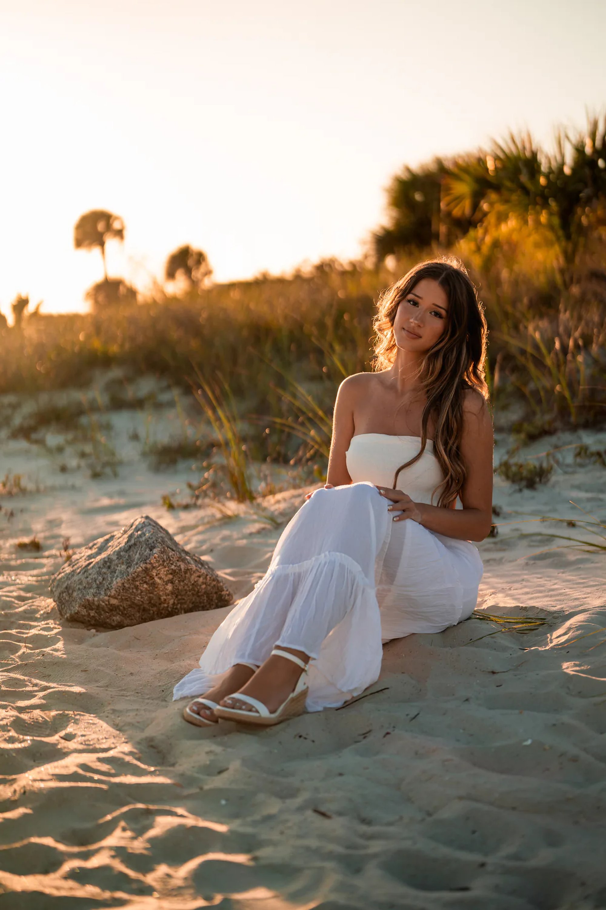 Young woman in a white dress seated on the sand at sunset.