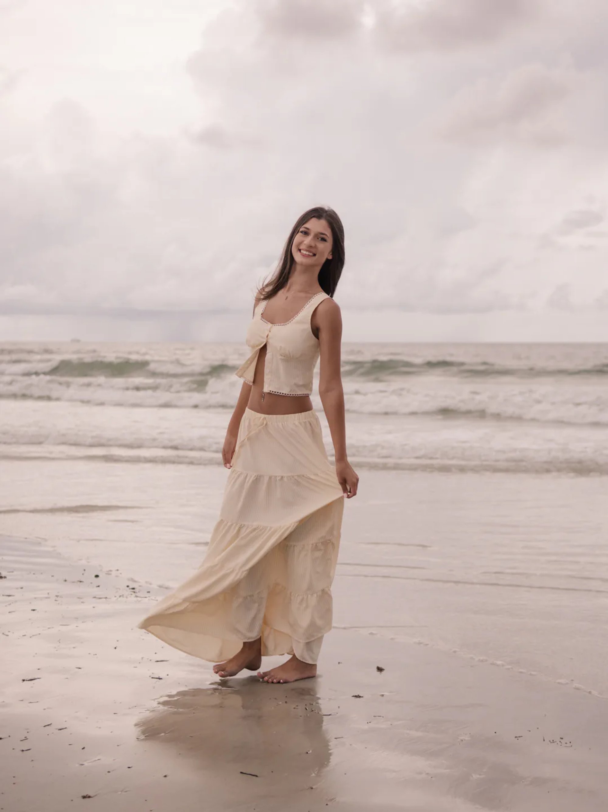 Ponce Inlet senior portraits, Mia standing barefoot in the water wearing a cream two-piece outfit, natural candid photo.