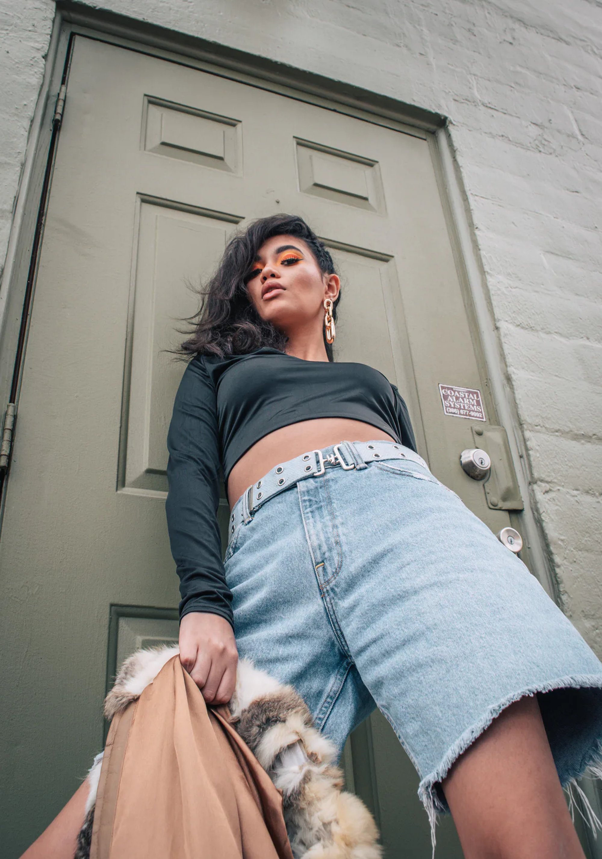 Low-angle portrait of a woman in a crop top and denim shorts leaning against a door.
