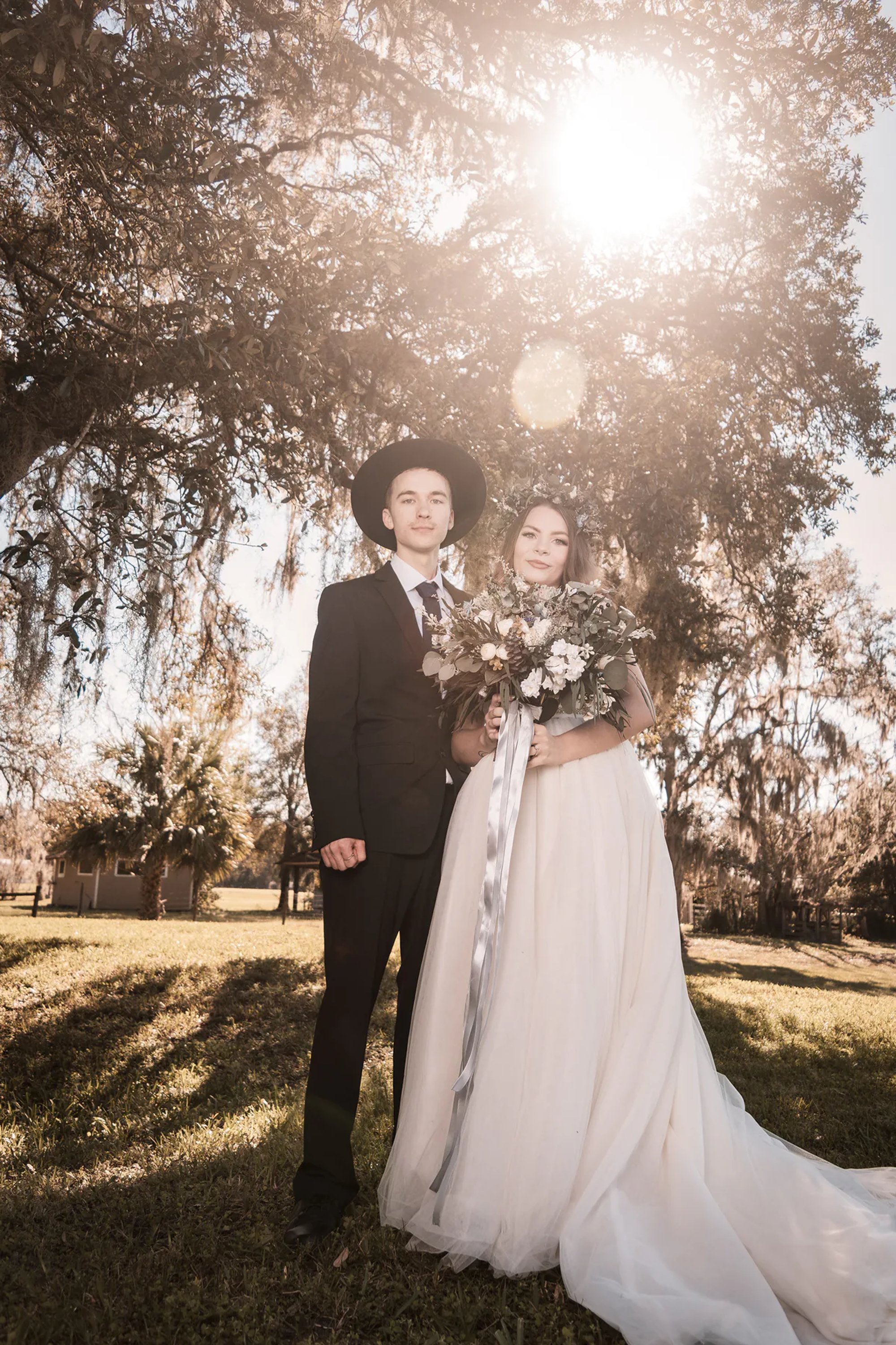 Newlyweds standing beneath oak trees with sunlight behind them.