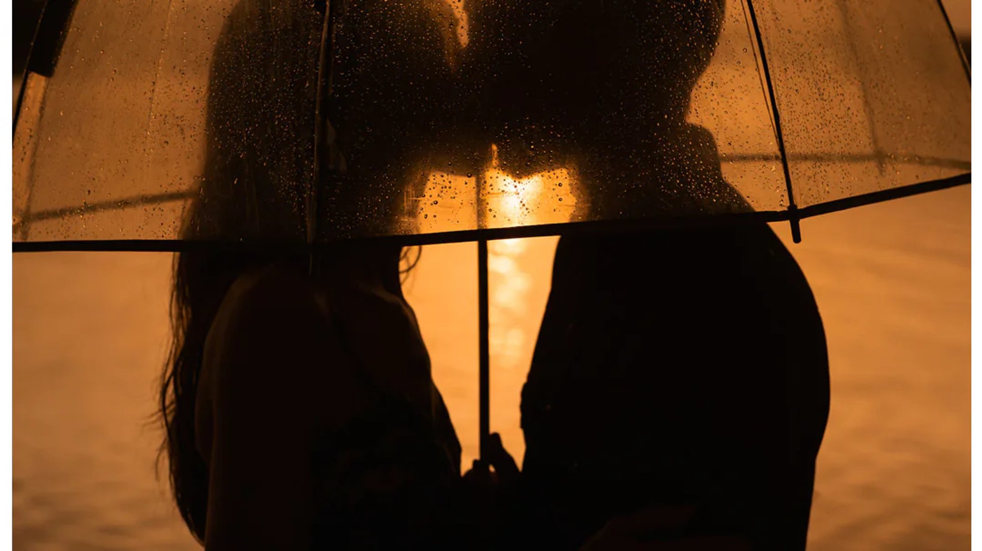 Couple kissing behind a rain-speckled umbrella at sunset.
