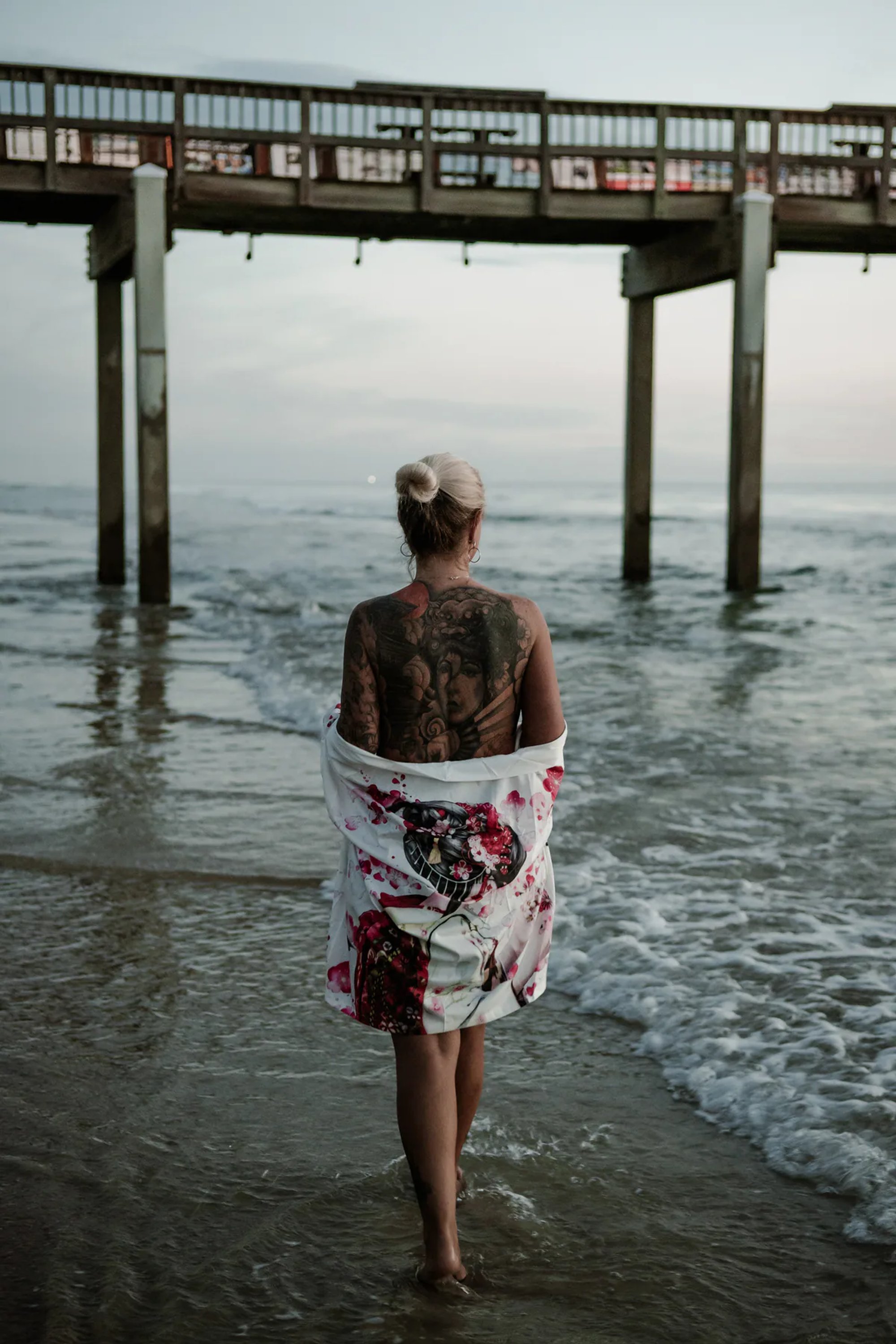 Tattooed woman holding a floral wrap while facing the ocean beneath a pier.