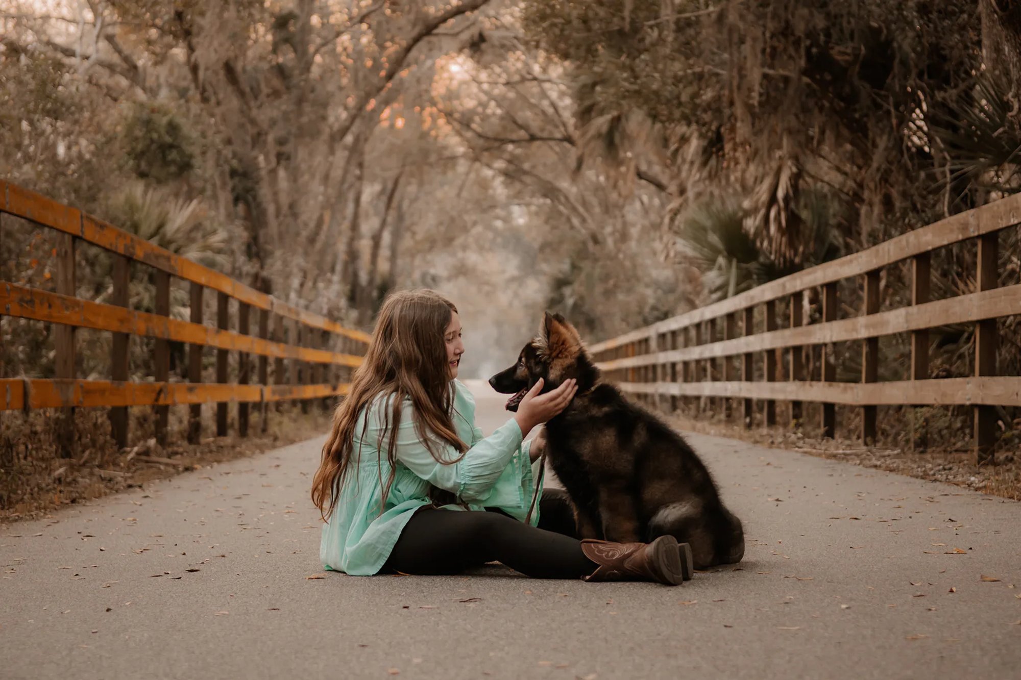 Young woman facing her seated black dog on a tree-lined road.